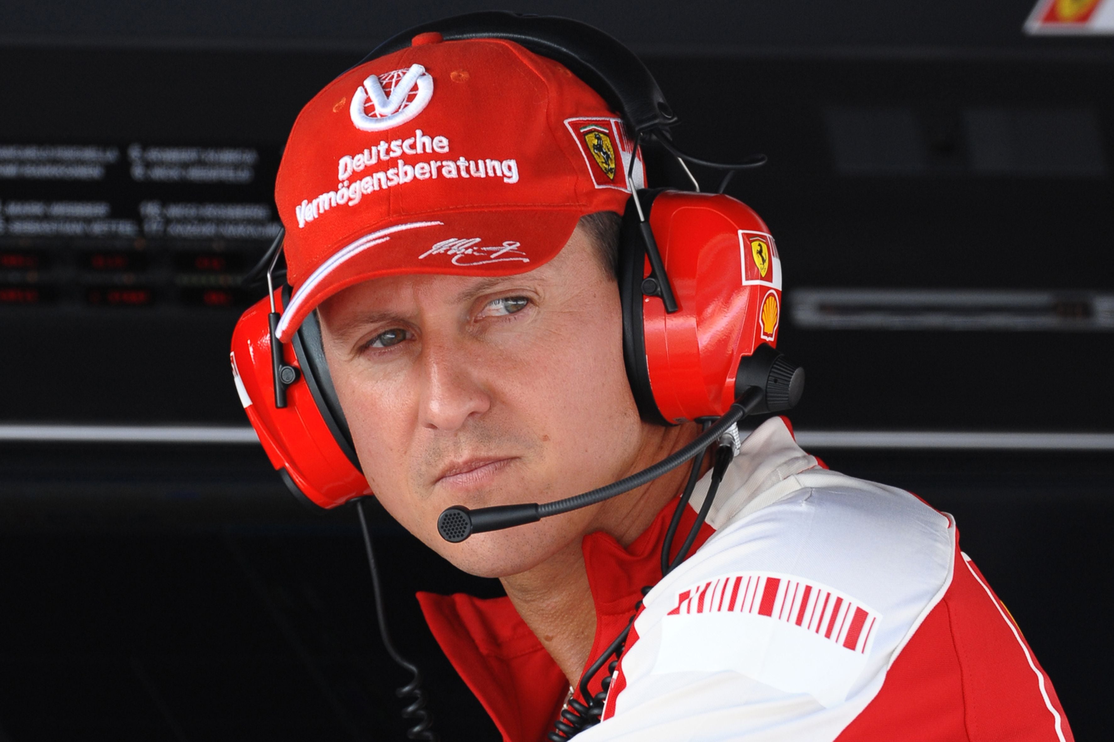 Ein Mann mit roter Ferrari-Kappe und Headset sitzt konzentriert an einer Boxengasse.
