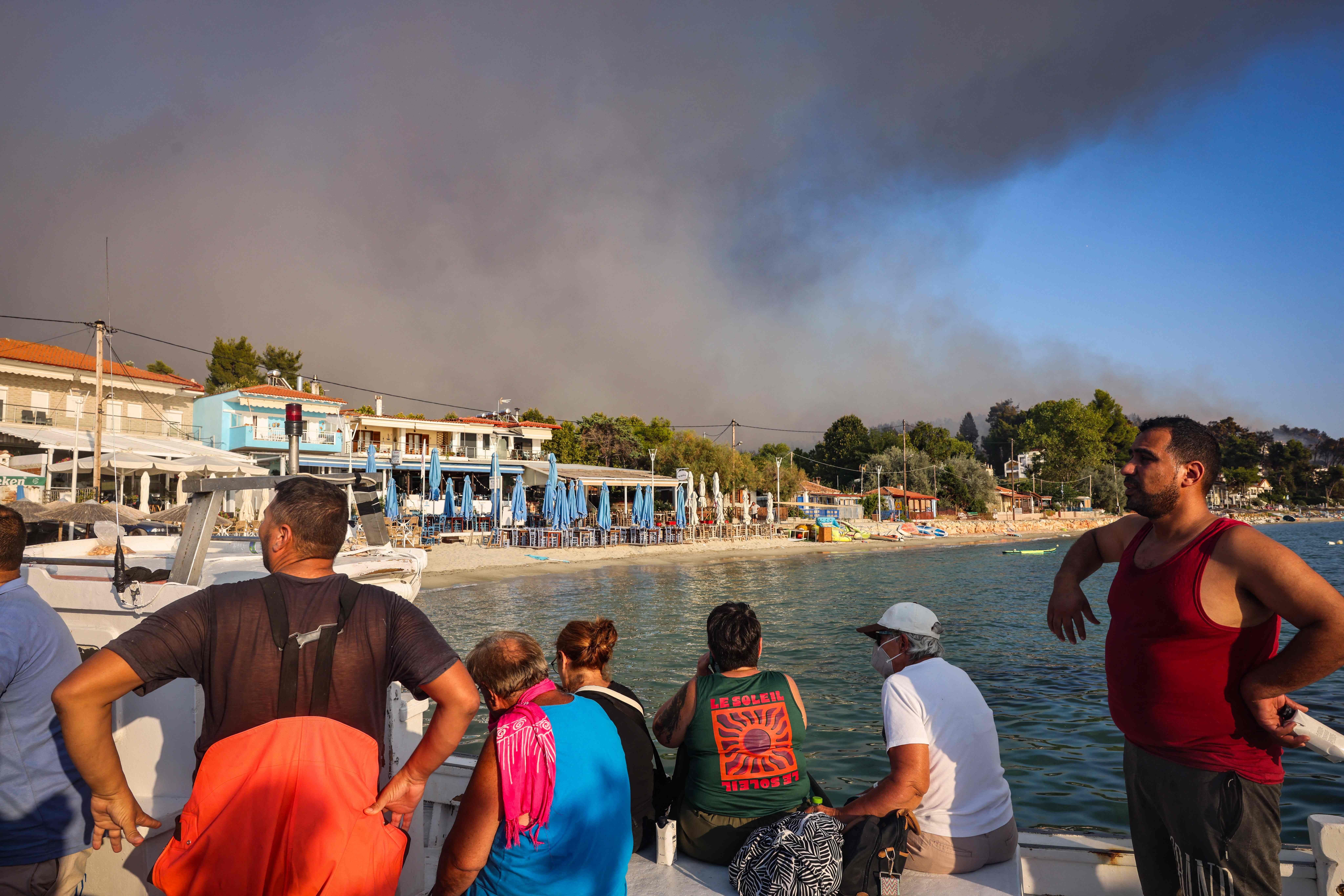 Menschen fliehen per Boot vor einem nahenden Waldbrand, Rauch steigt am Himmel bei Siviri auf.