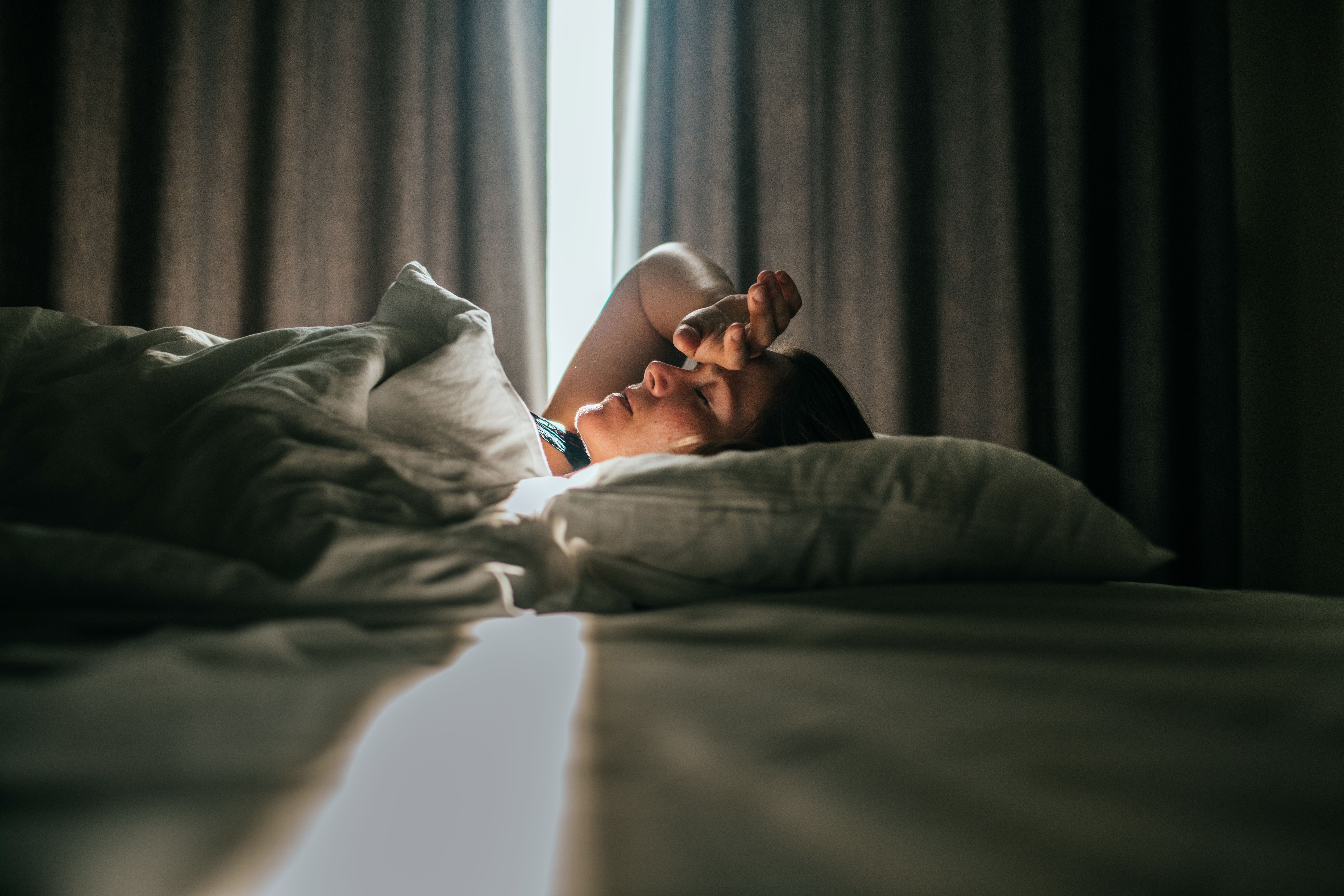 Frau liegt entspannt im Bett, Sonnenlicht fällt durch die Vorhänge, Hand an der Stirn.