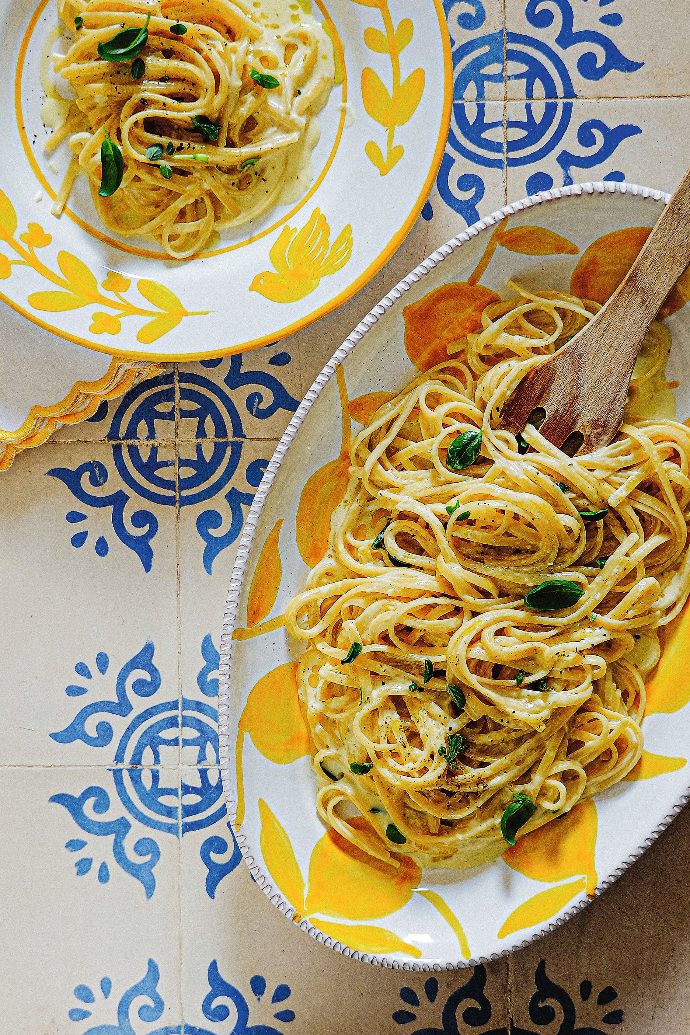 Spaghetti mit Sahnesoße und Basilikum auf buntem Geschirr neben Holzlöffel auf gemusterten Fliesen.