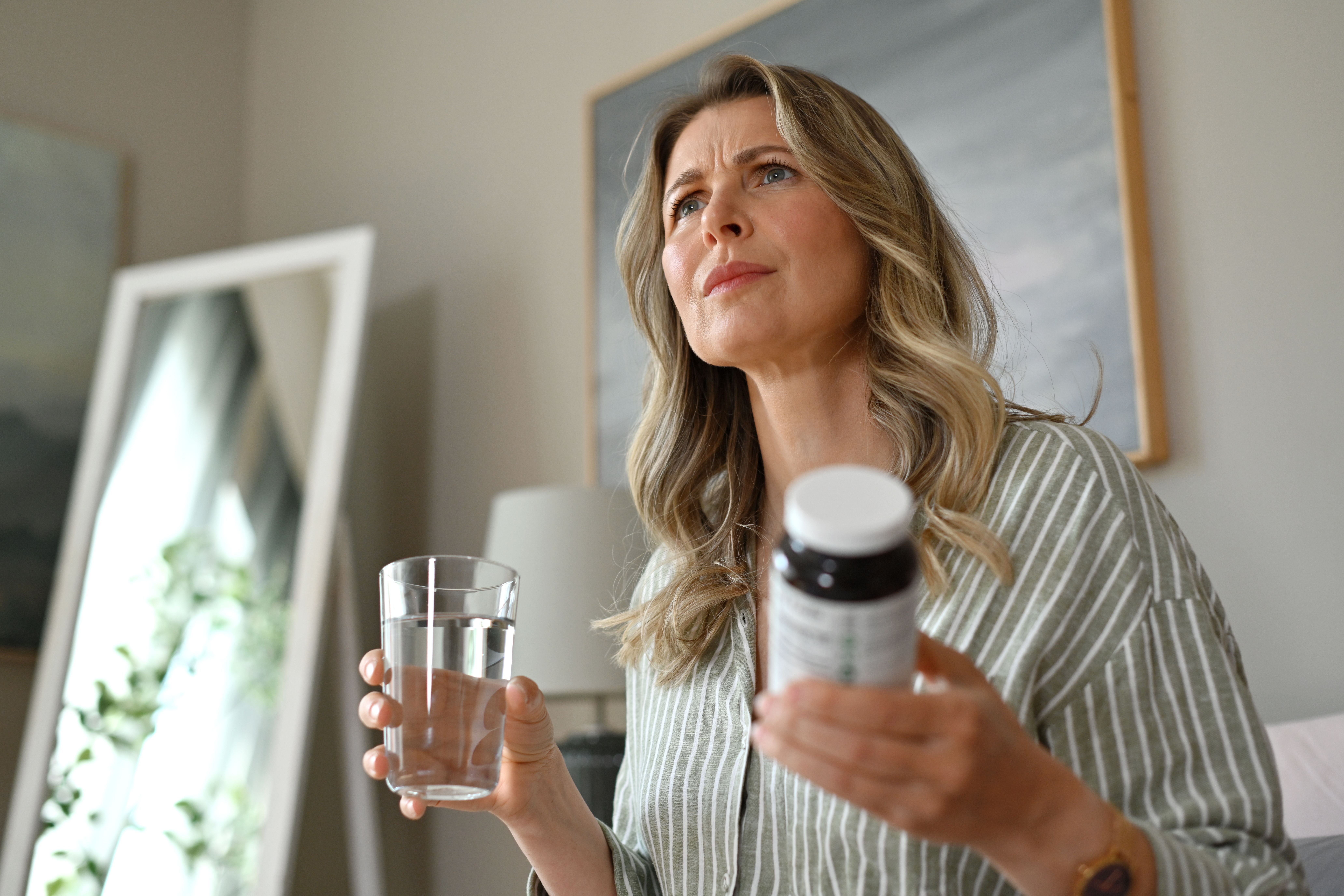 Frau hält ein Glas Wasser und eine Pillendose im Schlafzimmer und schaut nachdenklich.