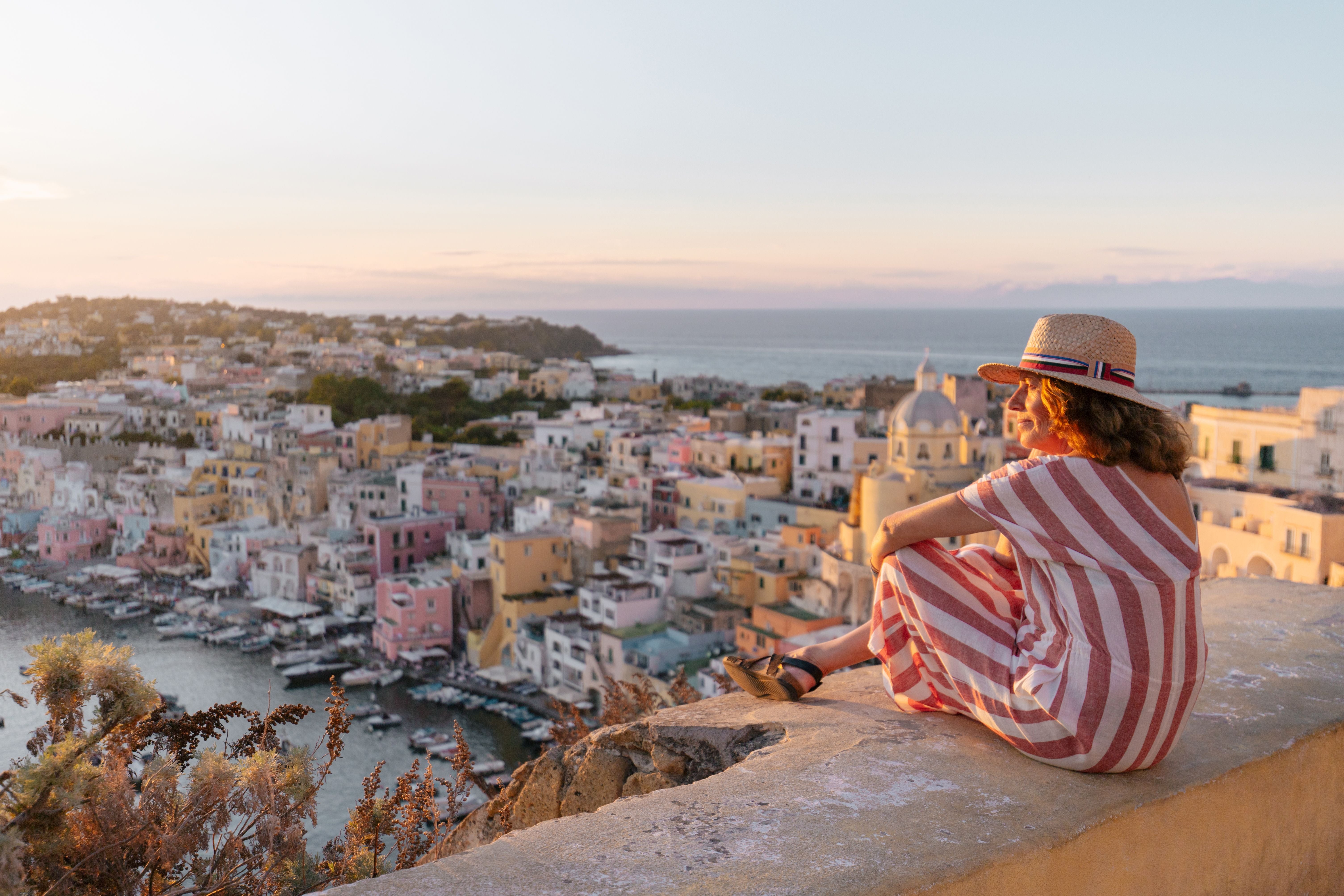 Frau in gestreiftem Kleid sitzt bei Sonnenuntergang auf einer Mauer und blickt auf Procida und das Meer.
