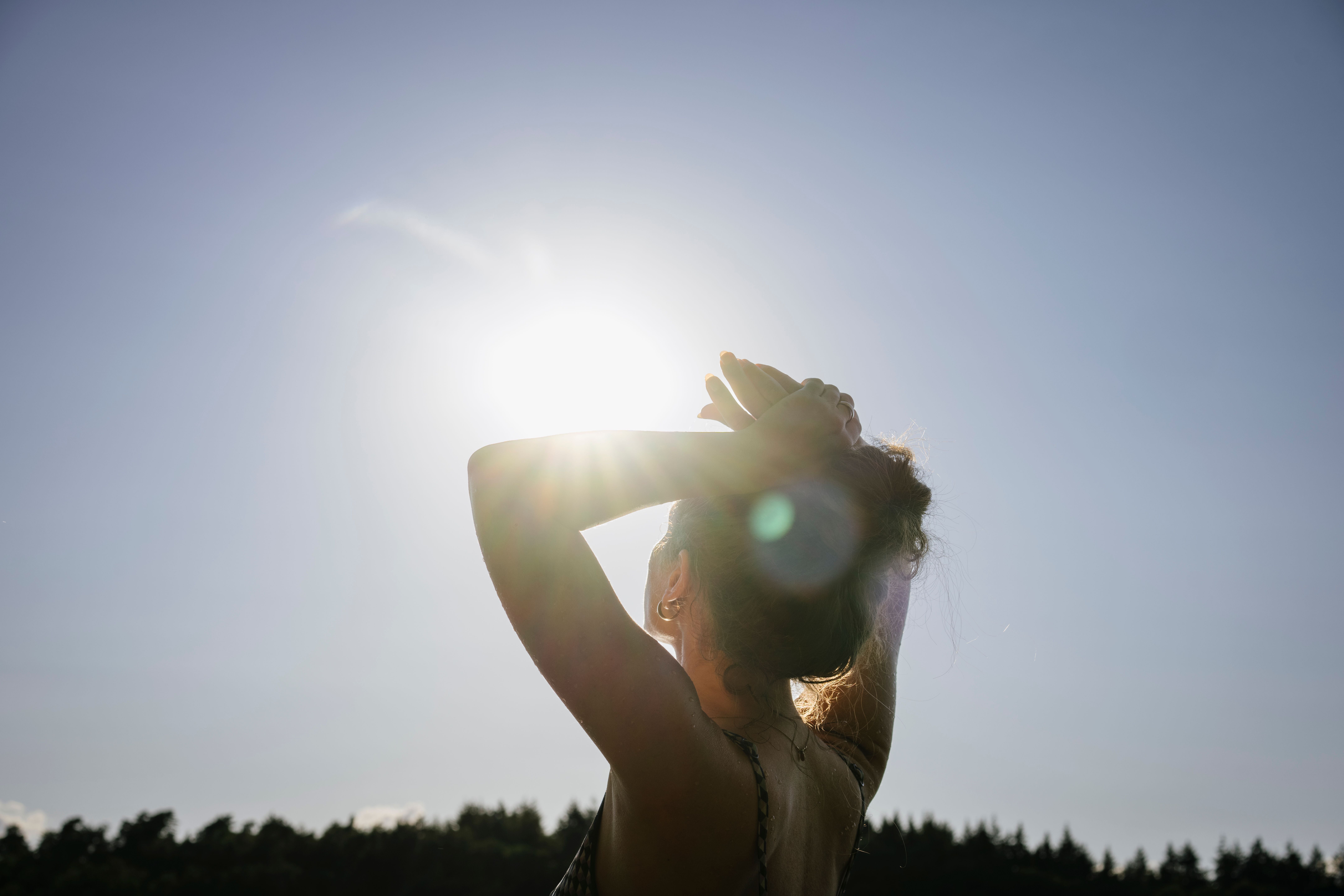 Frau von hinten, hält die Arme über dem Kopf und blickt in die Sonne am klaren Himmel.