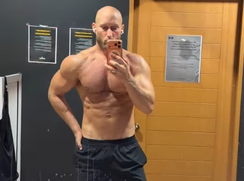 Ein muskulöser Mann macht ein Selfie vor einem Spiegel im Fitnessstudio.