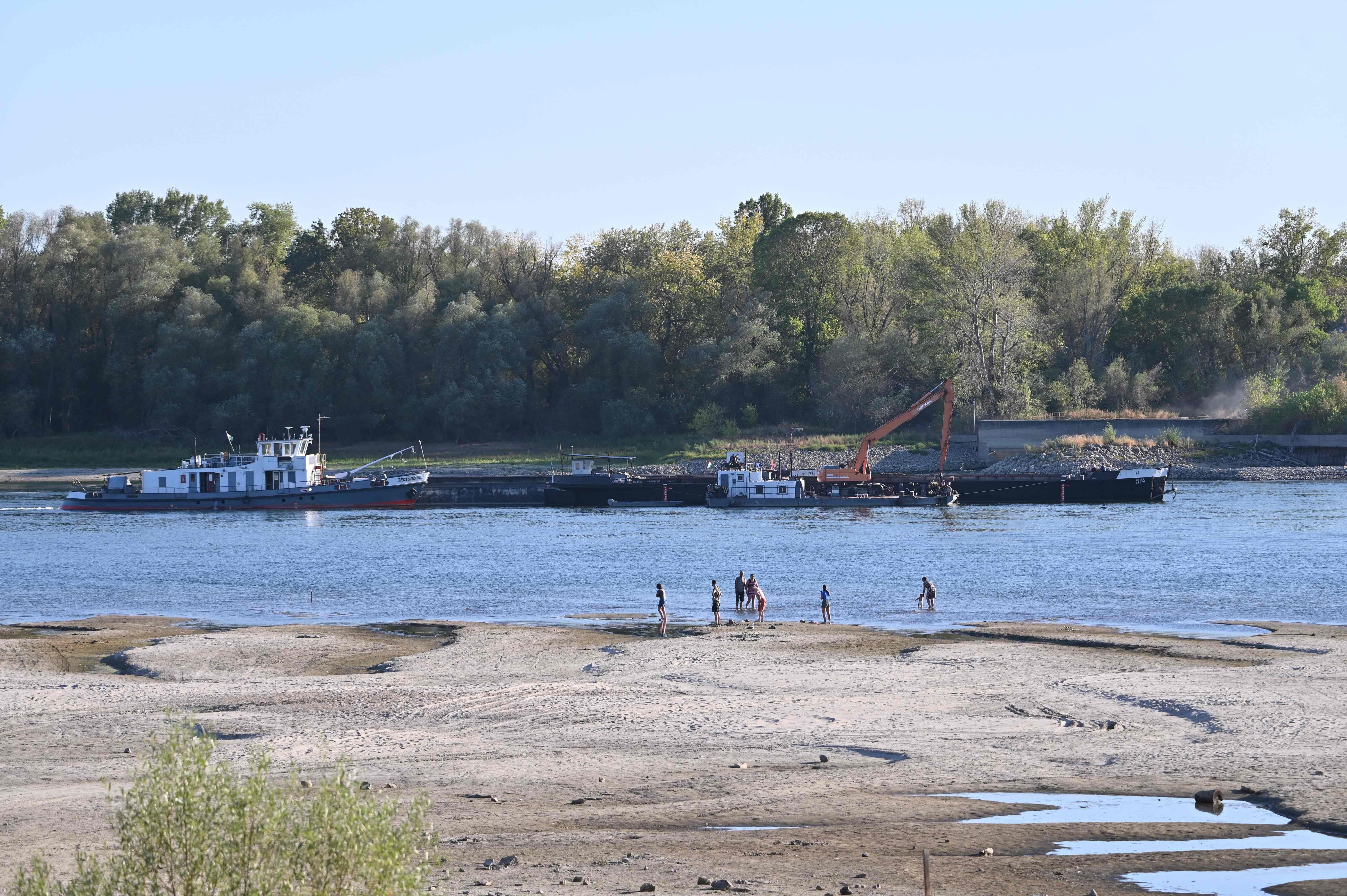 Schubschiff mit Felsen und Bagger auf der Donau; Menschen waten am niedrigen Ufer im Vordergrund.