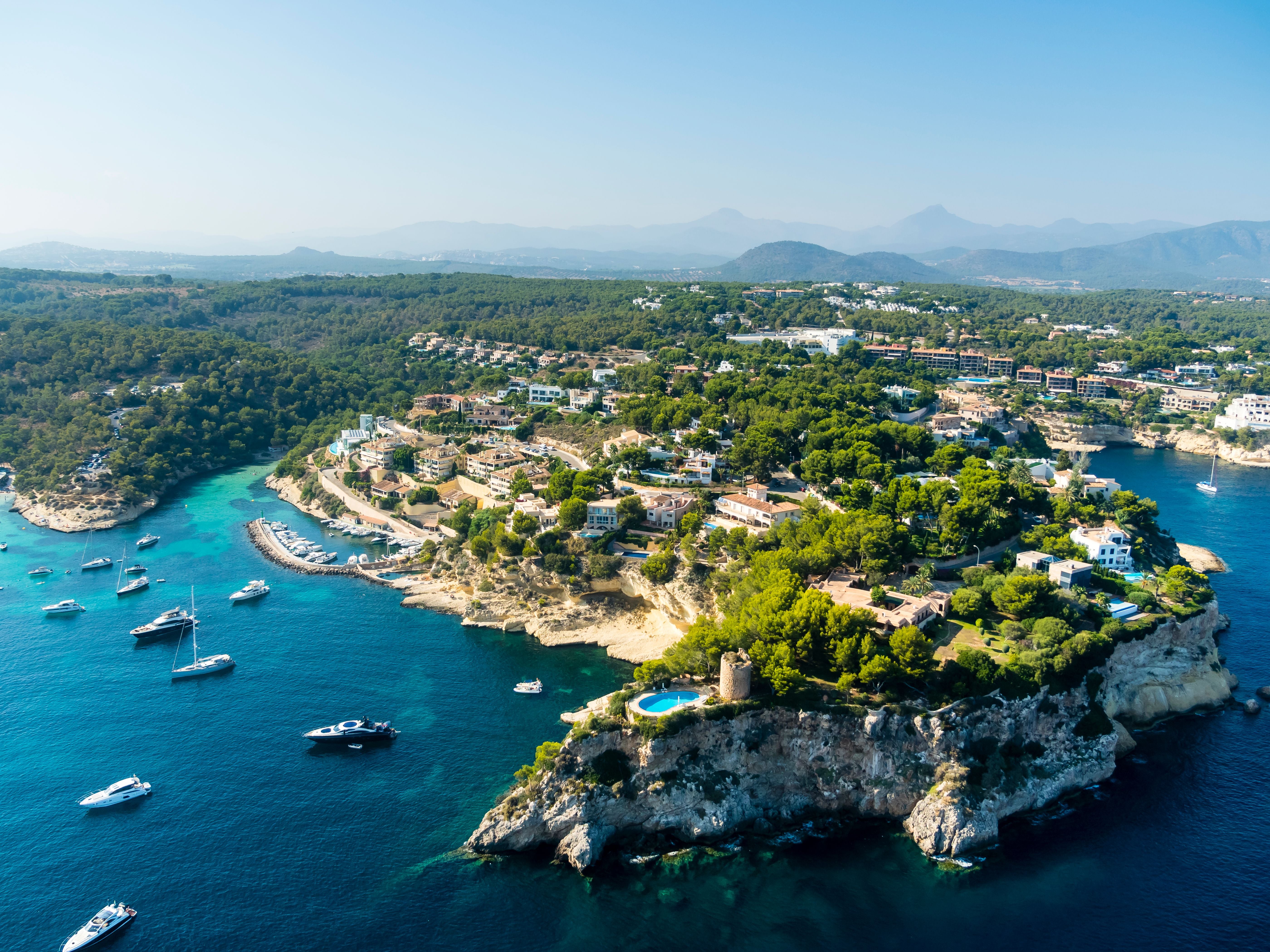 Luftaufnahme einer Küstenlandschaft mit Villen, Pinien und Yachten im türkisblauen Meer.