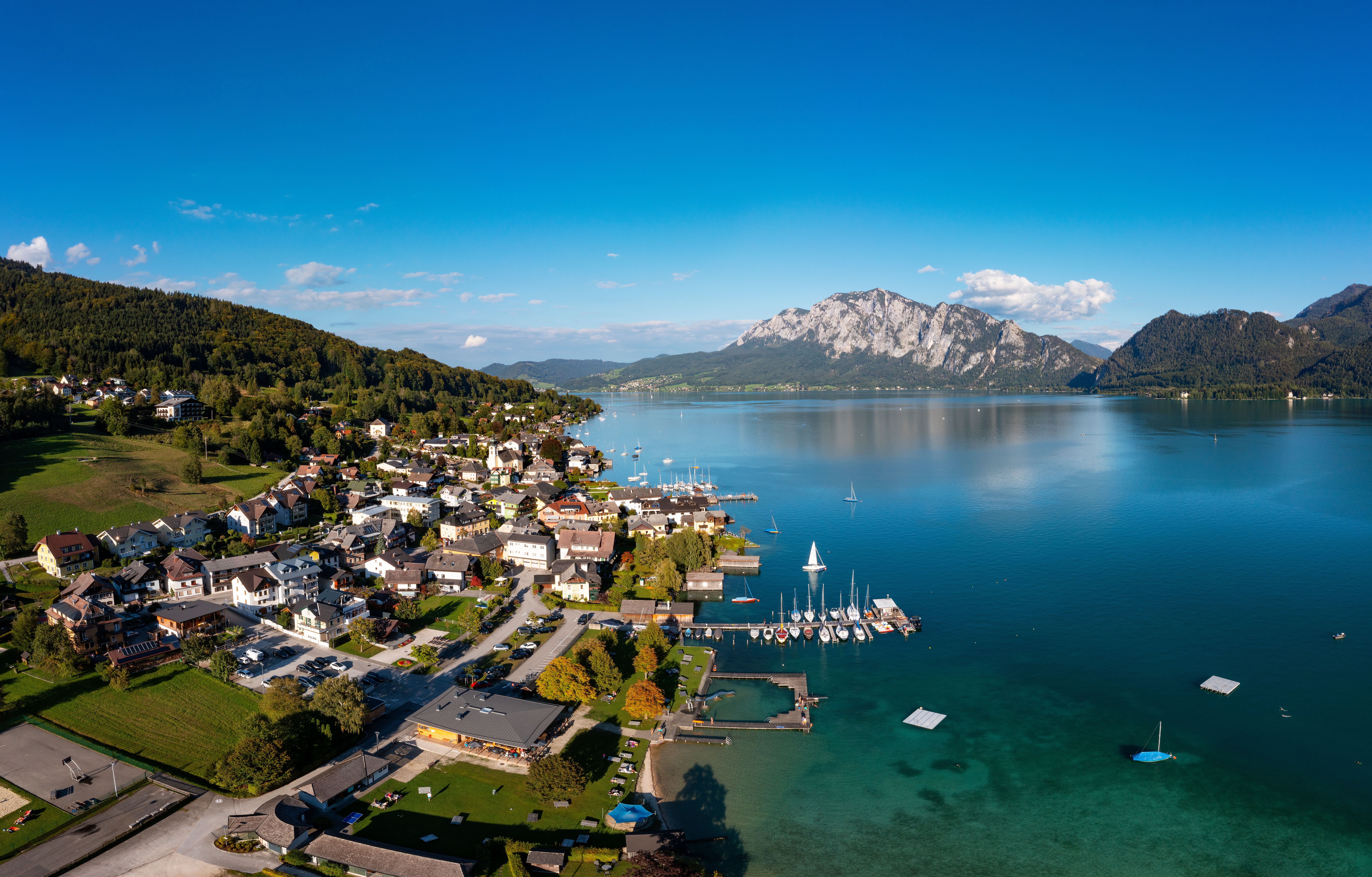 Luftaufnahme eines Dorfes am See mit Segelbooten und Bergen im Hintergrund an einem sonnigen Tag.