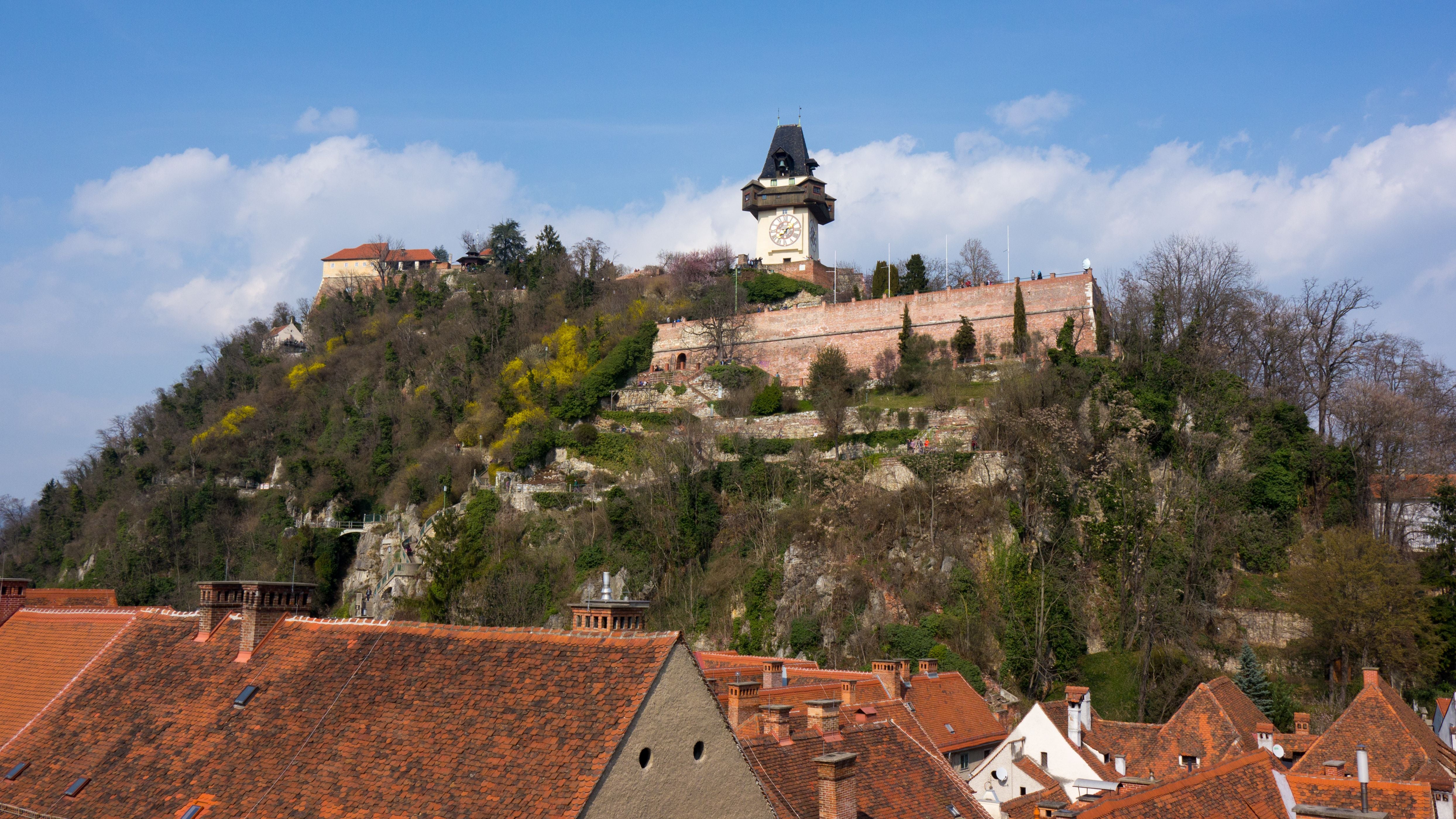 Der Schlossberg in Graz mit Uhrturm, umgeben von grüner Vegetation und roten Ziegeldächern.