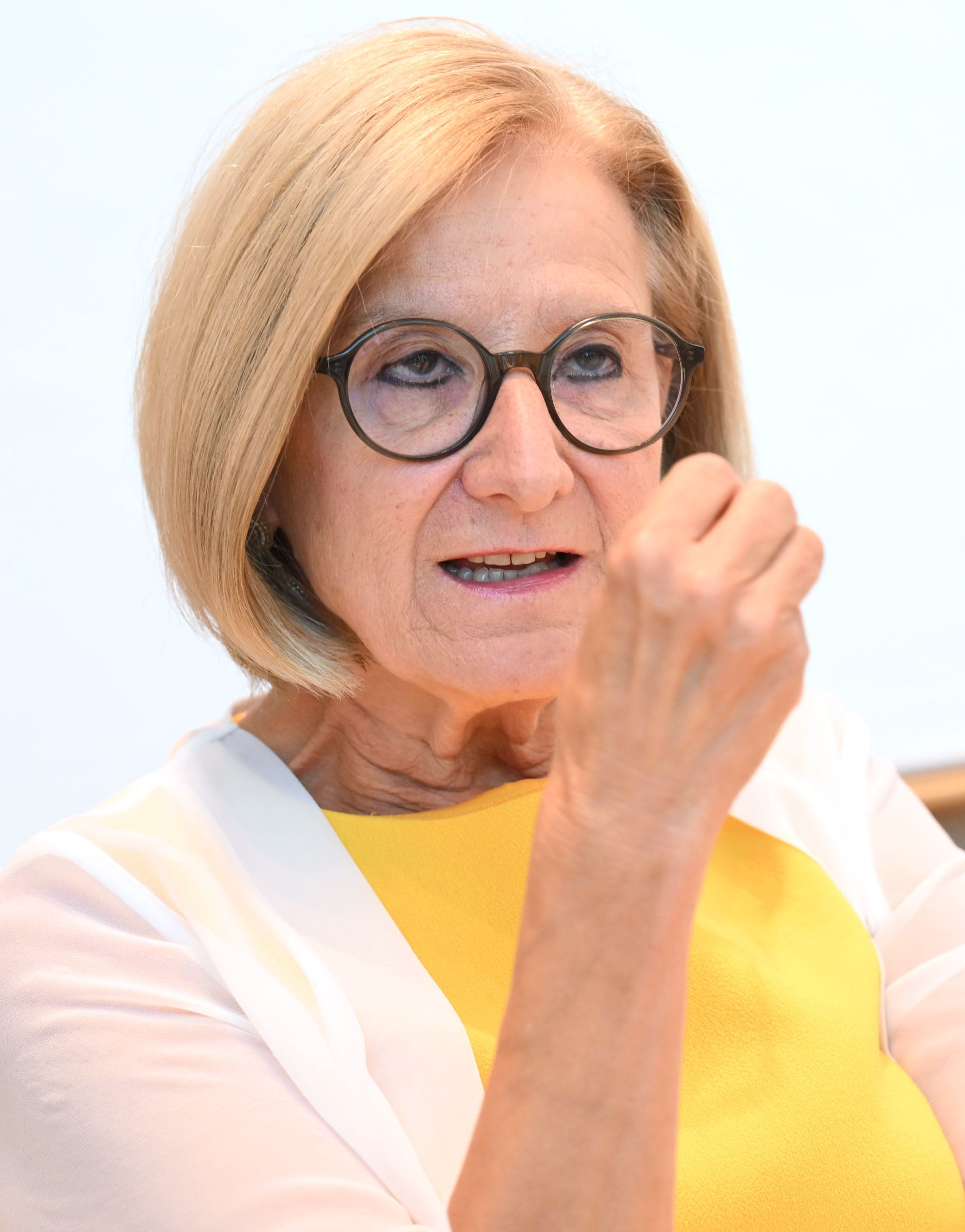 Ältere Frau mit blonden Haaren und Brille spricht in einem Interview, trägt gelbes Oberteil.
