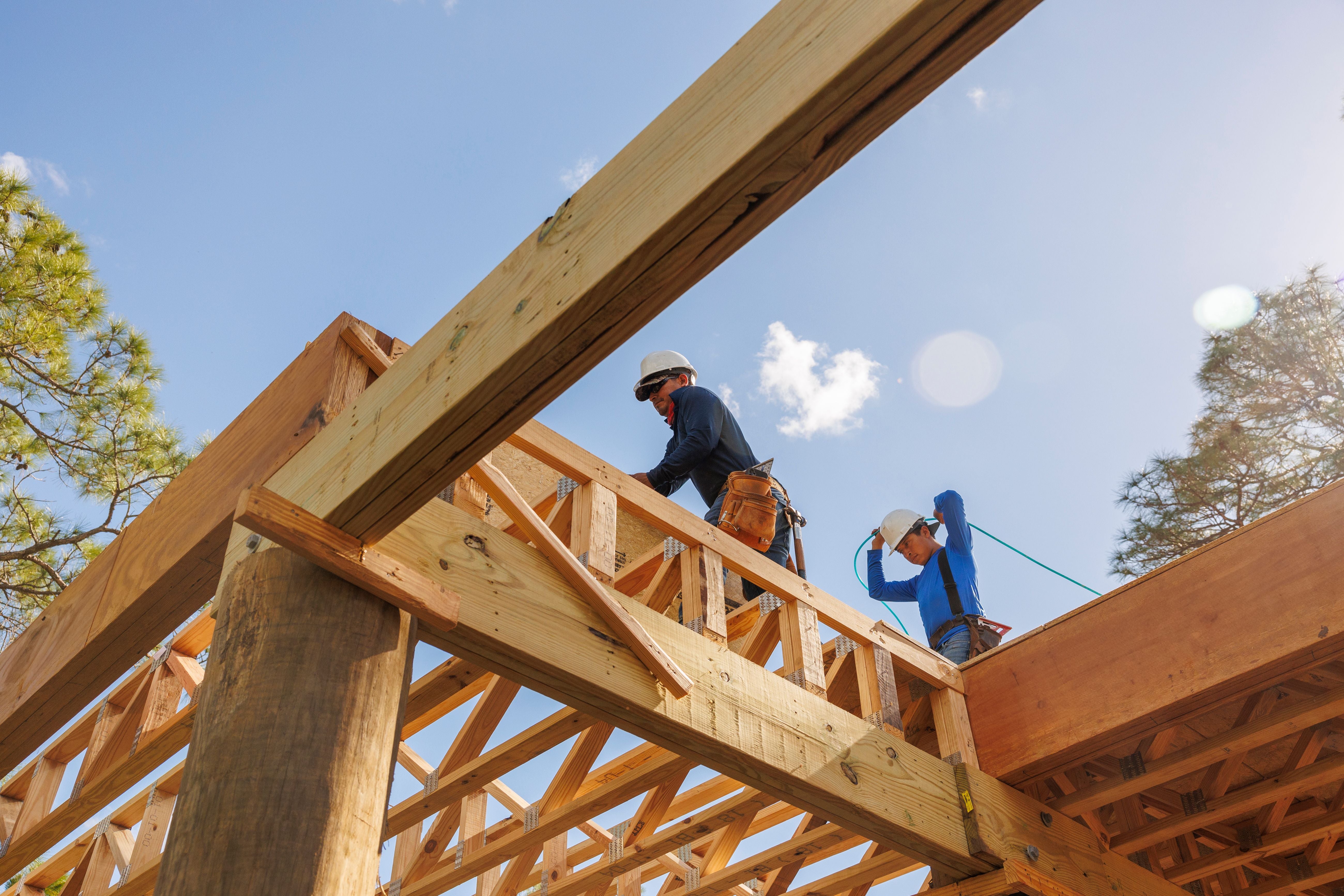 Zwei Bauarbeiter mit Helmen arbeiten auf einer Holzbaustelle unter blauem Himmel.