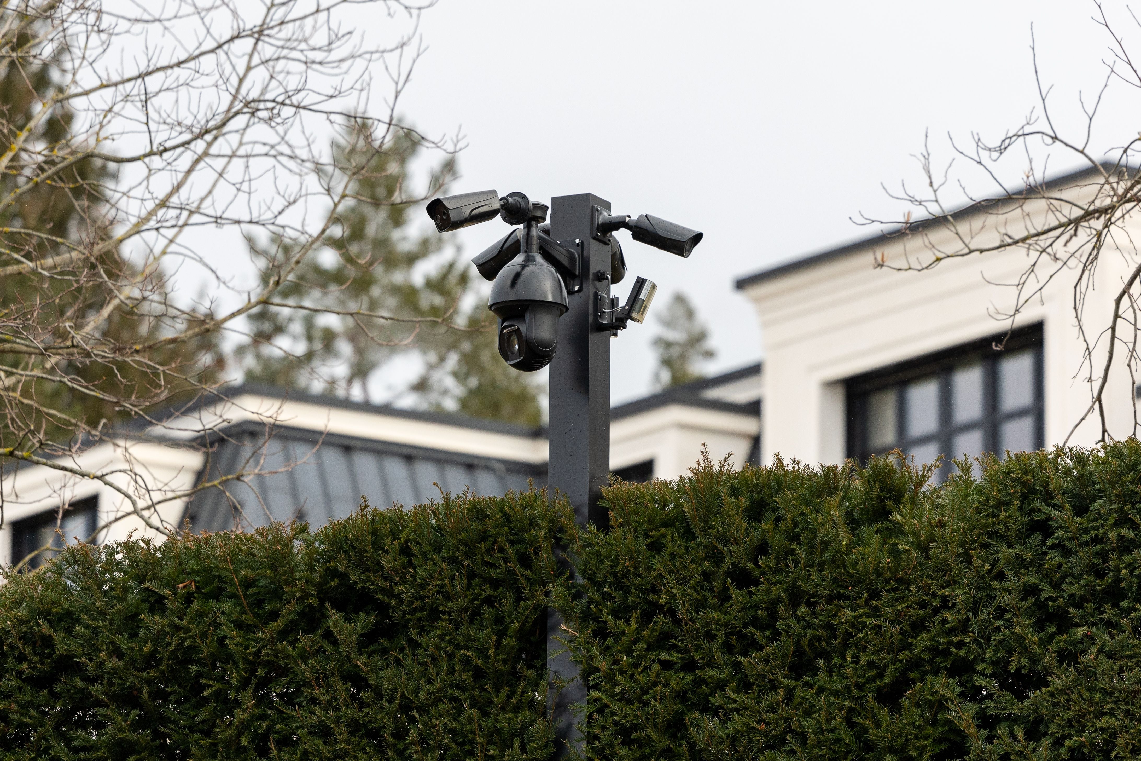 Mehrere Überwachungskameras auf einem Mast vor einer modernen Villa hinter einer hohen Hecke.