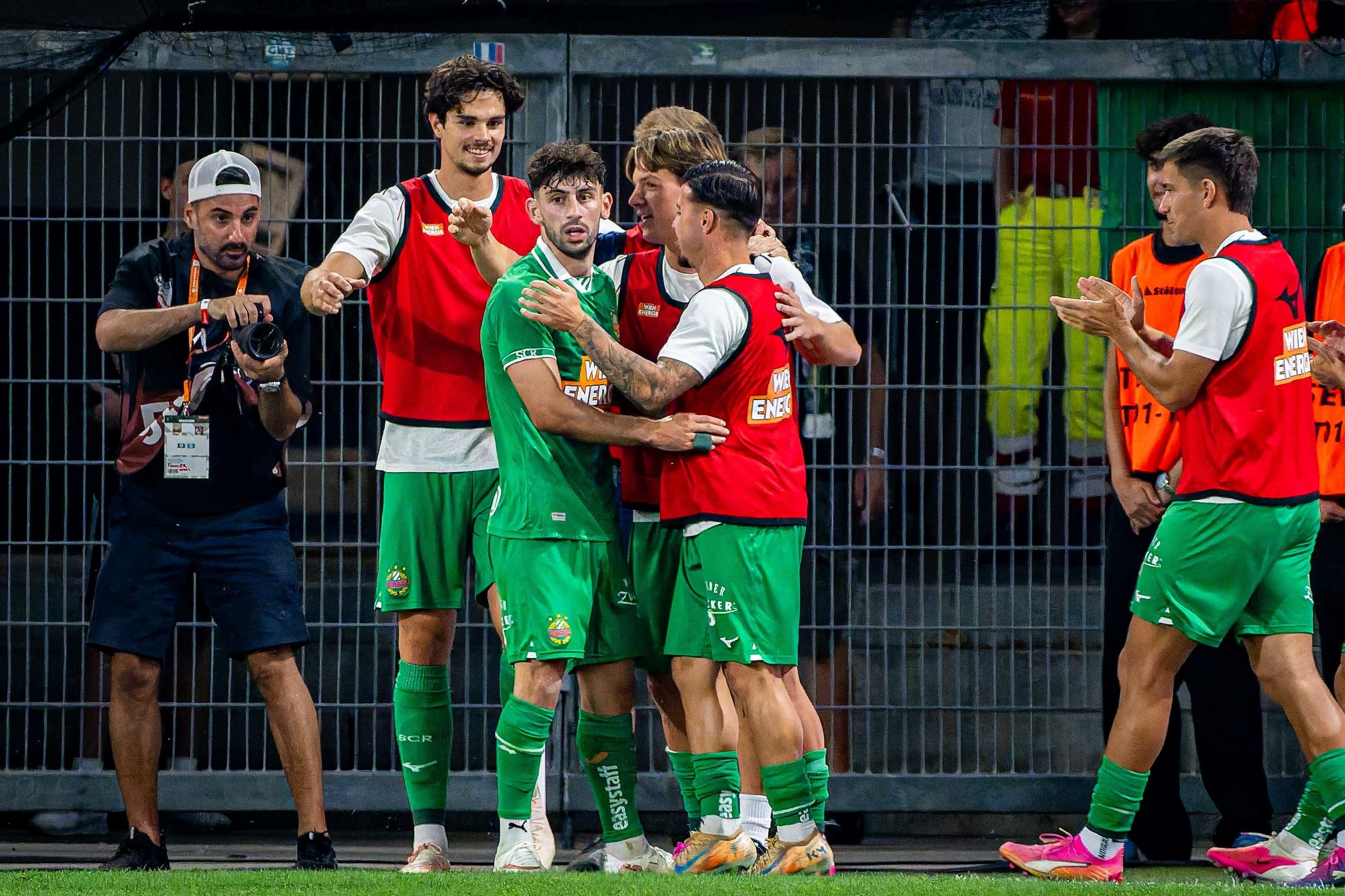 Mehrere Rapid-Spieler feiern jubelnd am Spielfeldrand, ein Fotograf hält die Szene fest.