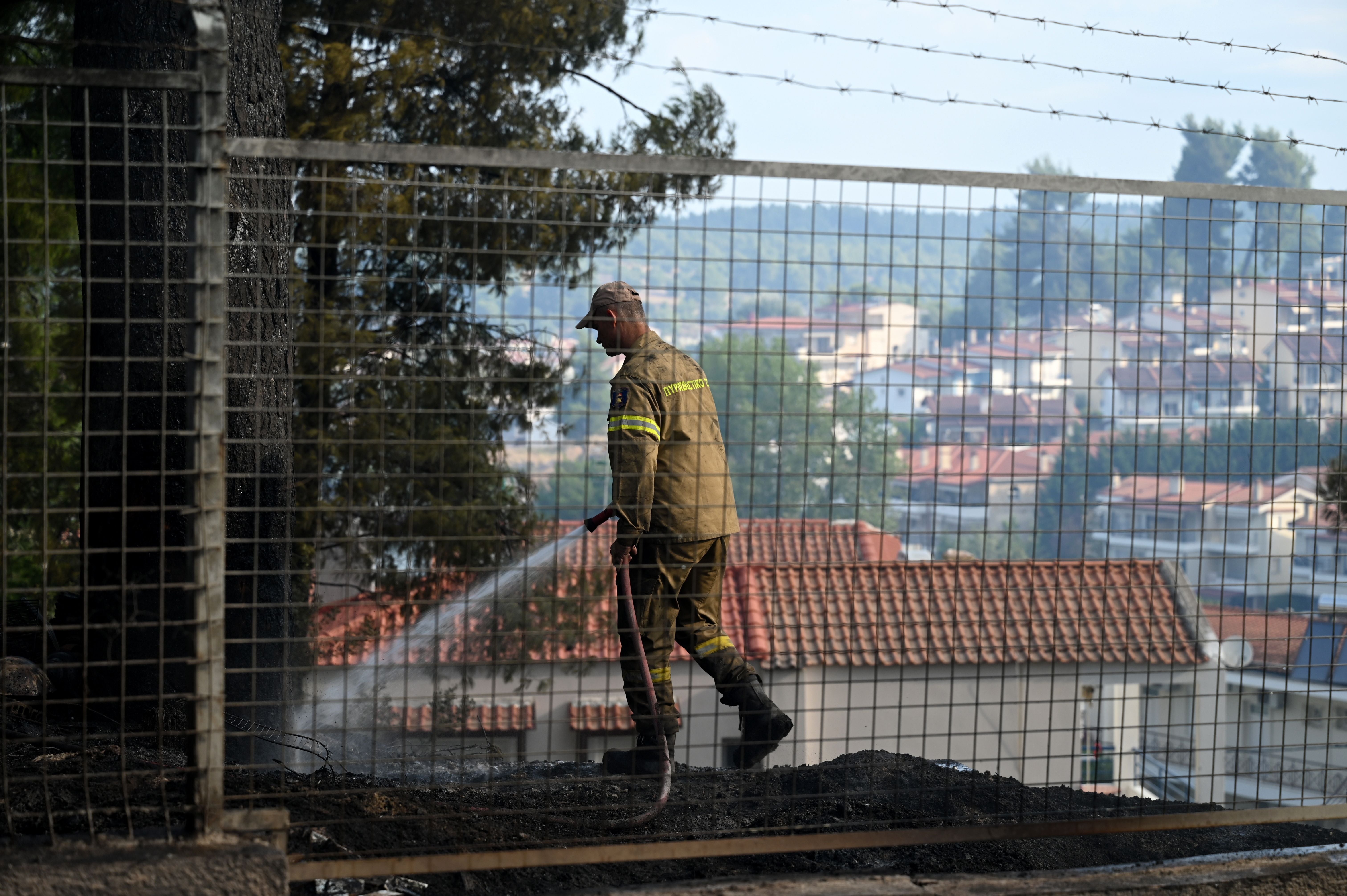 Ein Feuerwehrmann löscht mit einem Schlauch einen Brand in einem umzäunten, verbrannten Hof.