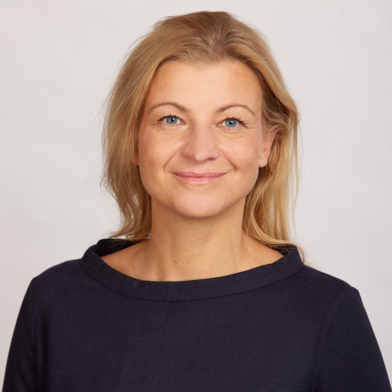 Frau mit blonden Haaren und dunkelblauem Oberteil vor hellem Hintergrund.