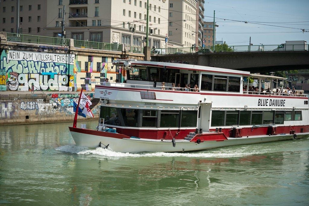 Ein Ausflugsschiff namens "Blue Danube" fährt auf einem Fluss an Graffiti und Gebäuden vorbei.