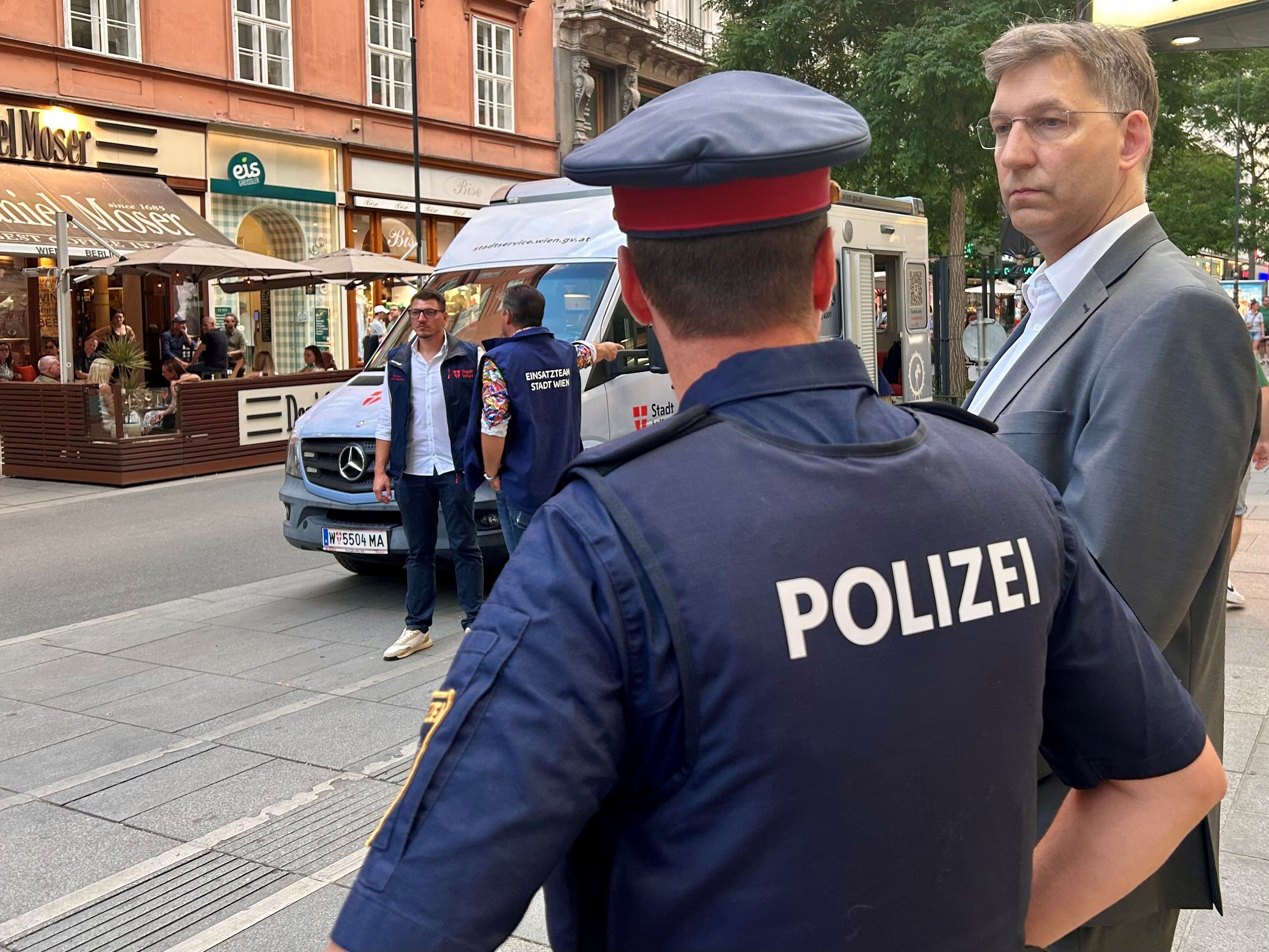 Ein Polizist steht auf einer belebten Straße neben mehreren Männern und einem Einsatzfahrzeug.