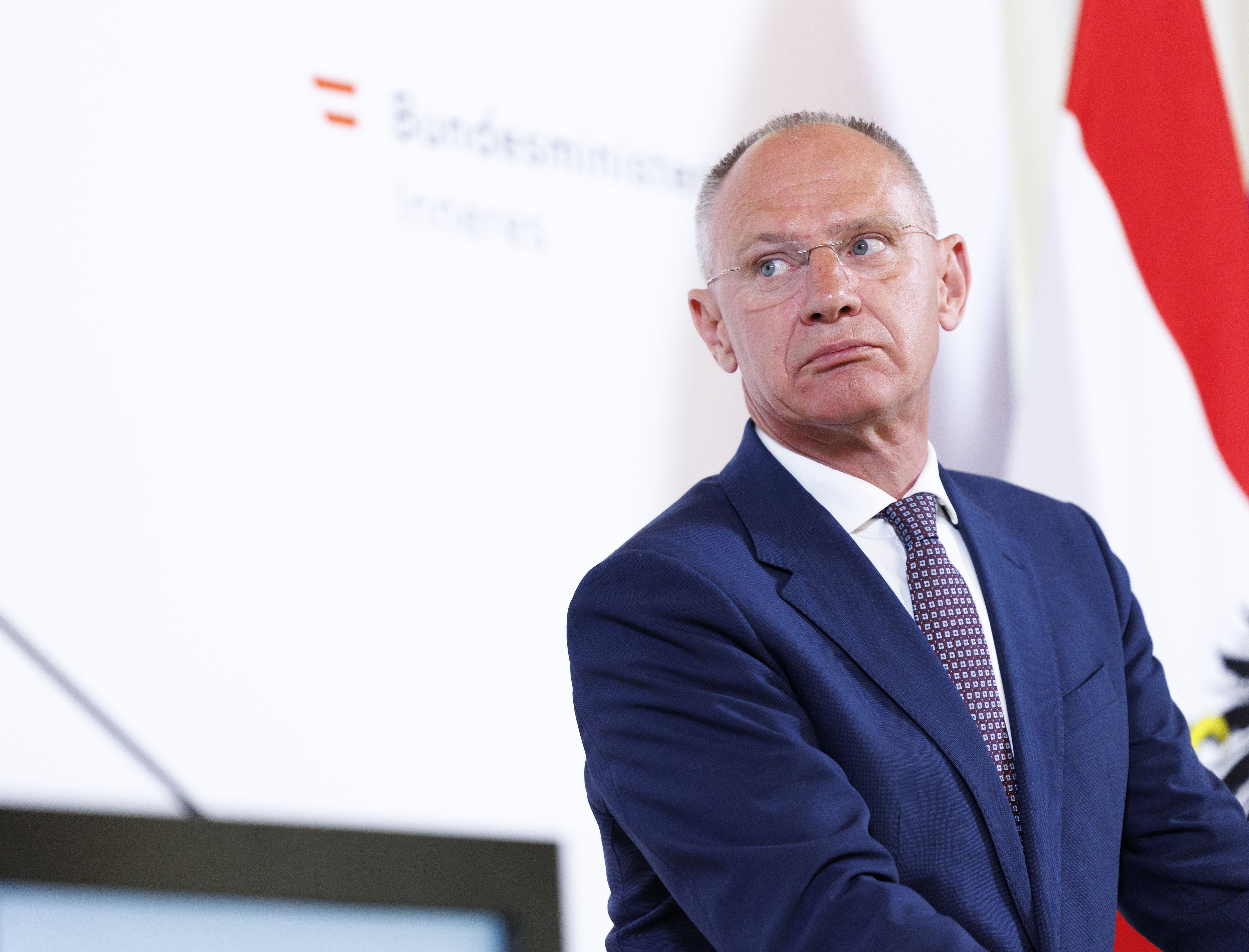 Ein Mann im blauen Anzug steht vor einer österreichischen Flagge und einem Ministeriumslogo.