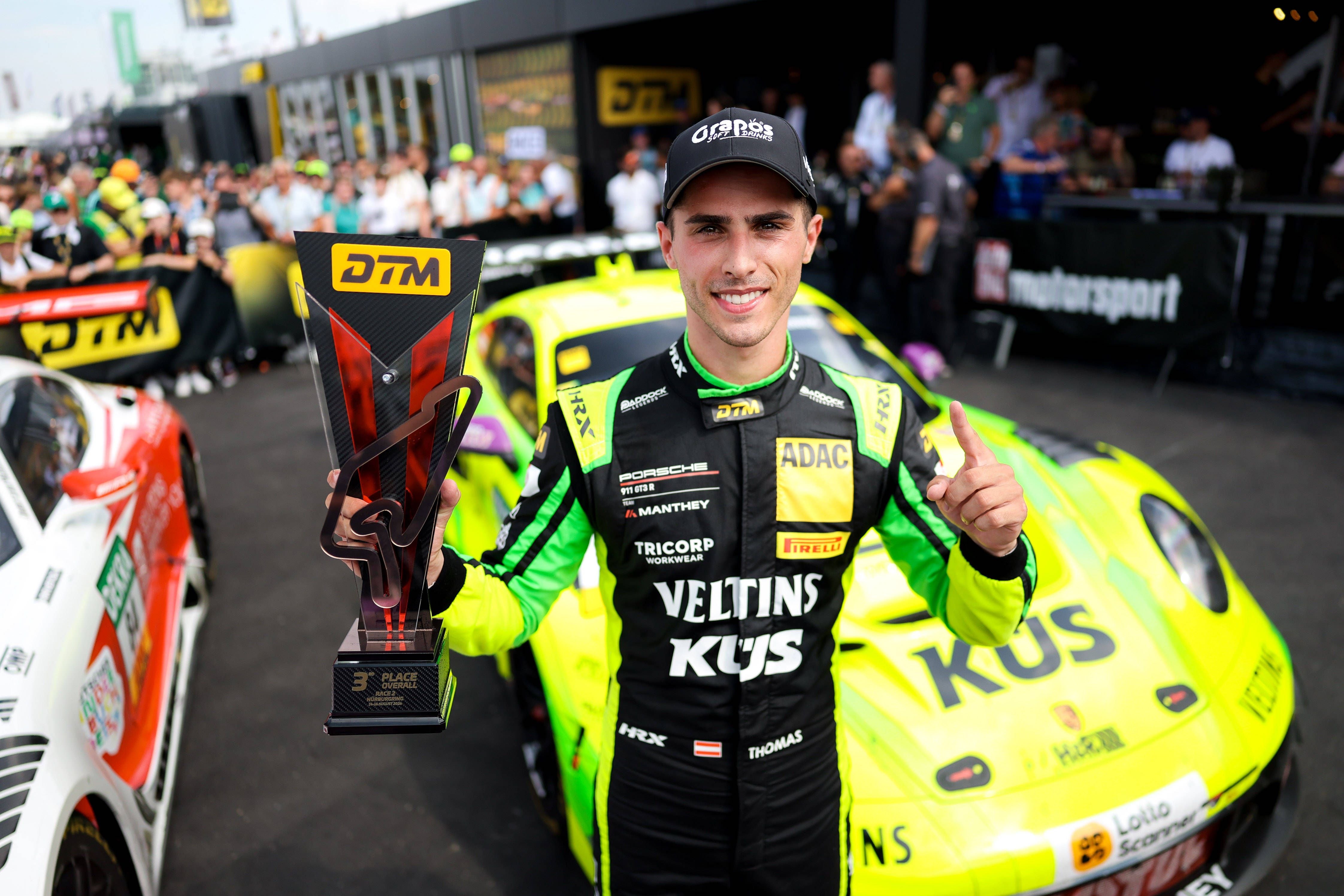 Thomas Preining hält eine DTM-Trophäe vor einem gelb-grünen Porsche 911 GT3 R am Nürburgring.