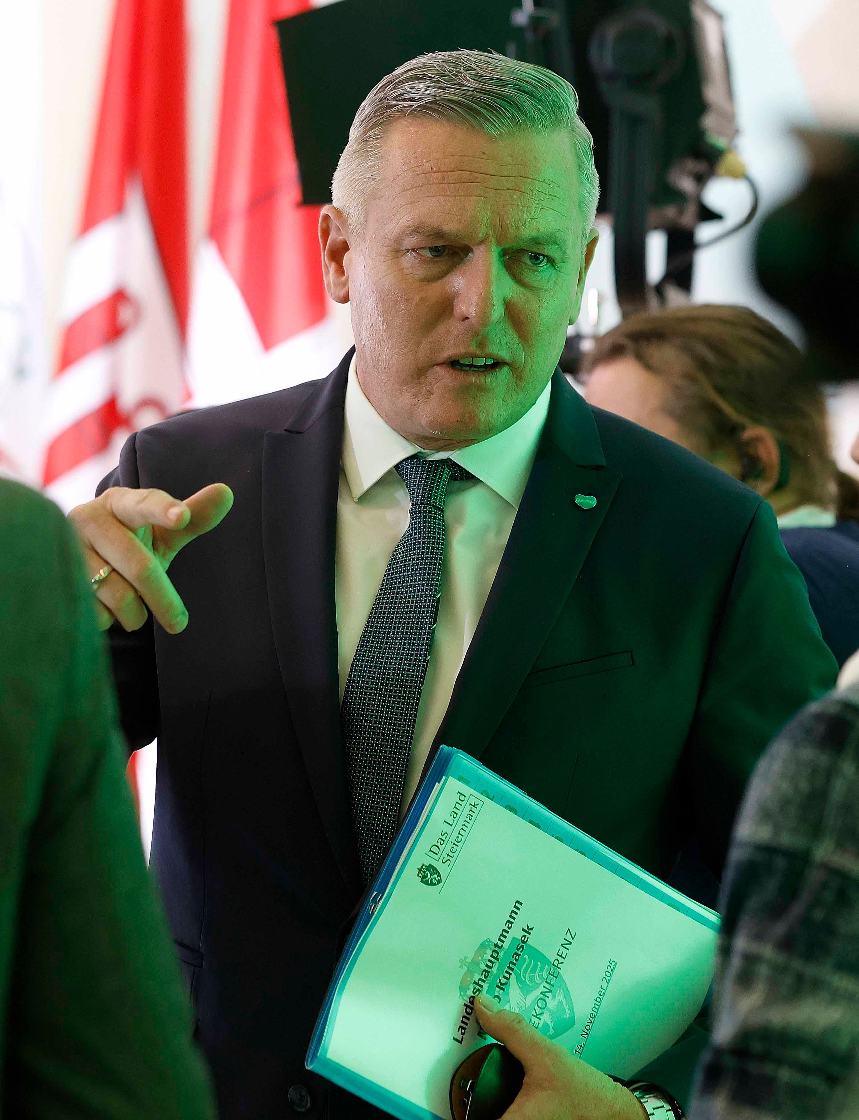 Ein Mann im Anzug hält blaue Unterlagen und spricht bei einer Pressekonferenz.