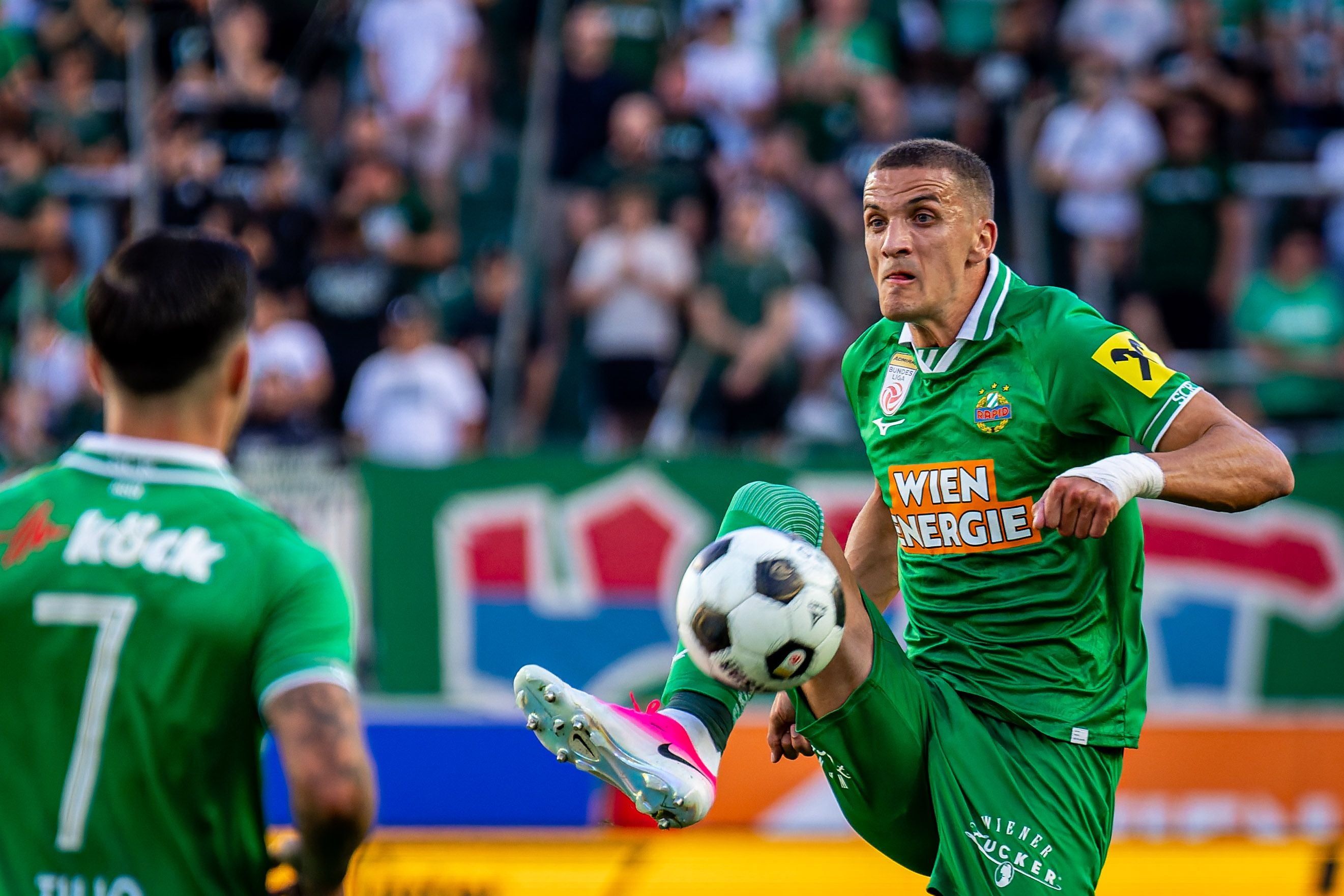 Fußballspieler von Rapid Wien schießt den Ball während eines Spiels, ein Mitspieler ist im Vordergrund.
