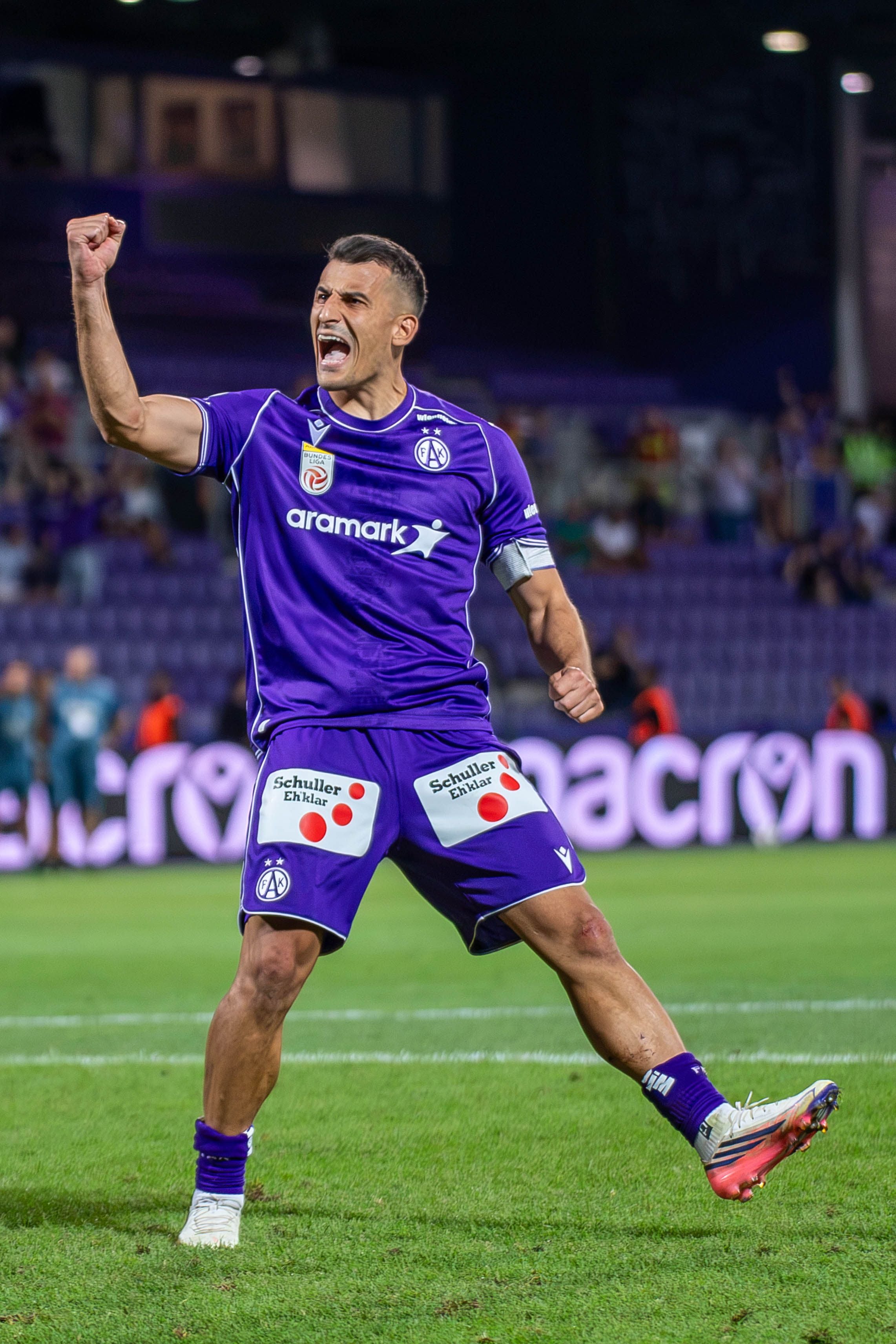 Ein Fußballspieler im violetten Trikot jubelt mit erhobener Faust auf dem Spielfeld.