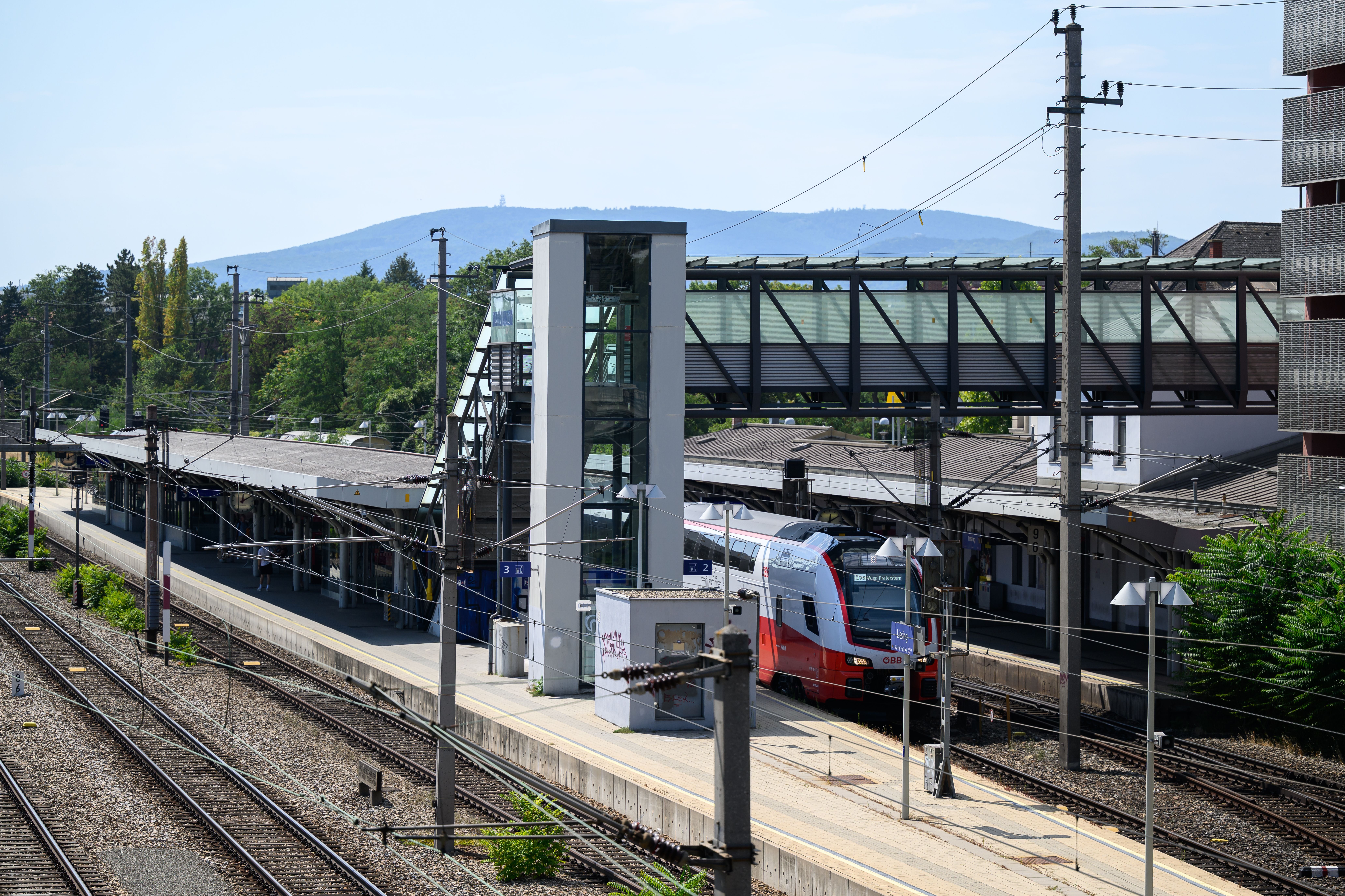 Bahnhof Wien-Liesing mit einem roten ÖBB-Zug und Fußgängerbrücke, sonniger Tag.