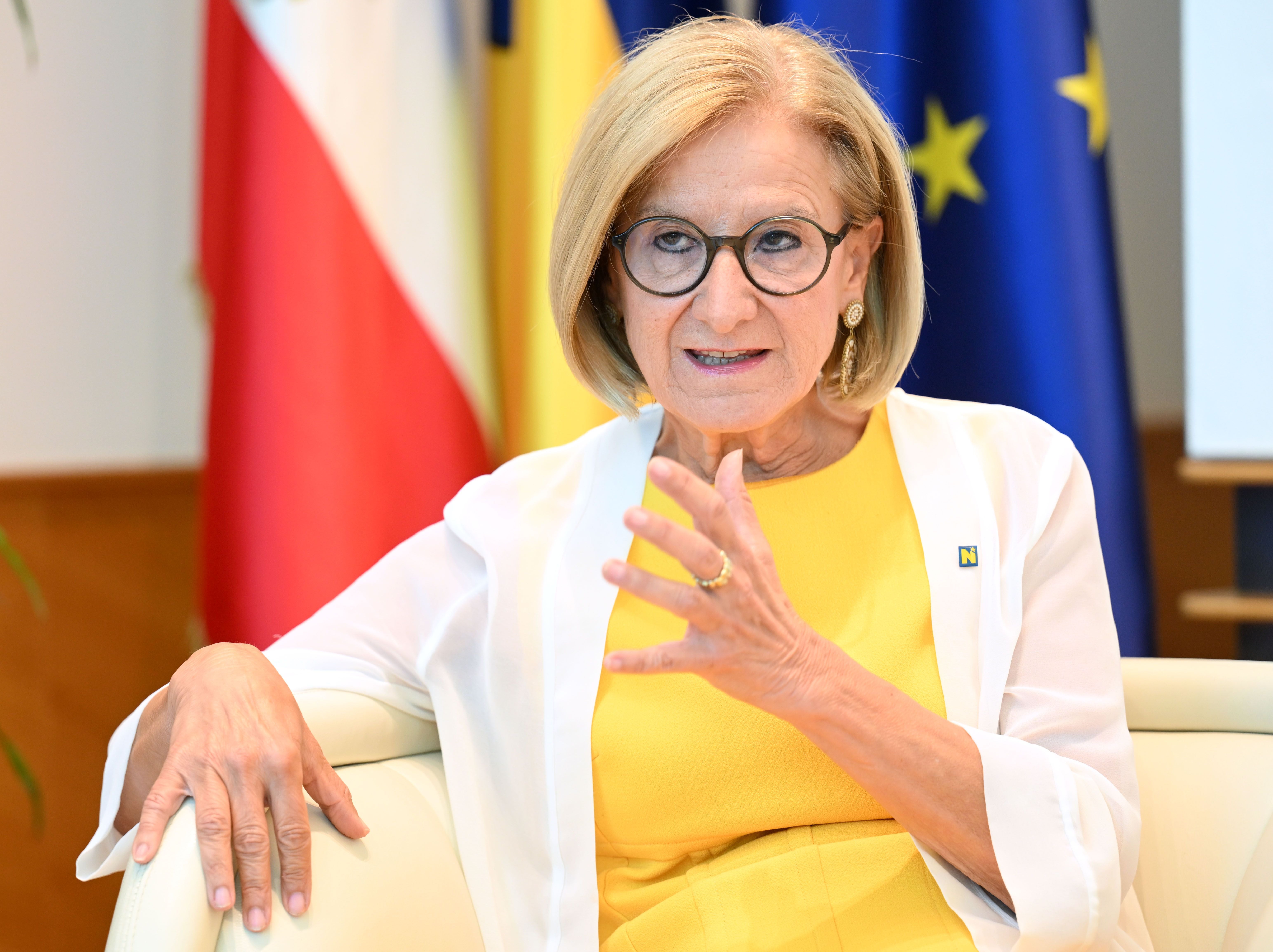 Frau mit Brille und gelbem Kleid spricht gestikulierend auf einem Sessel vor Fahnen.