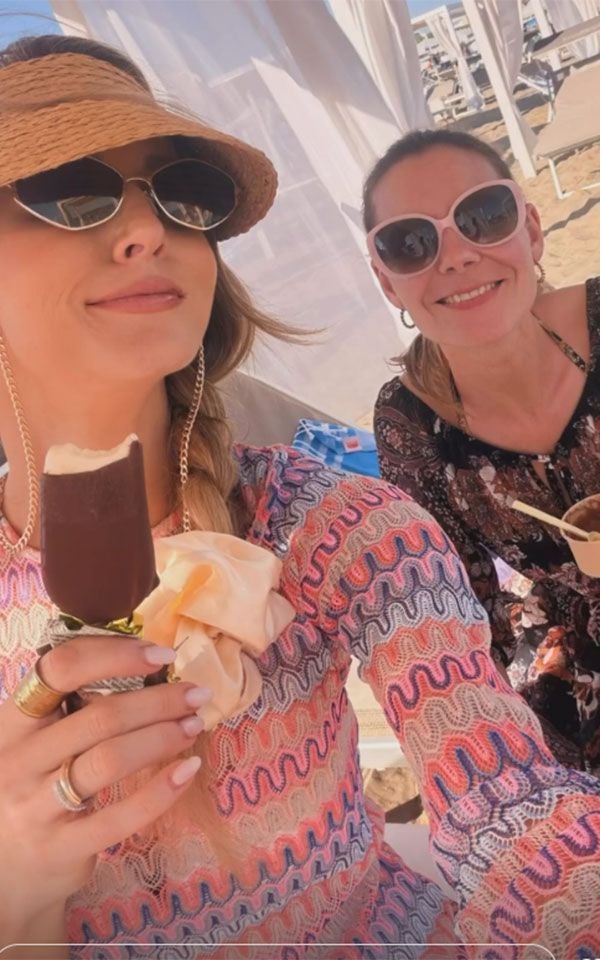 Zwei Frauen mit Sonnenbrillen genießen Eis am Strand und lächeln in die Kamera.