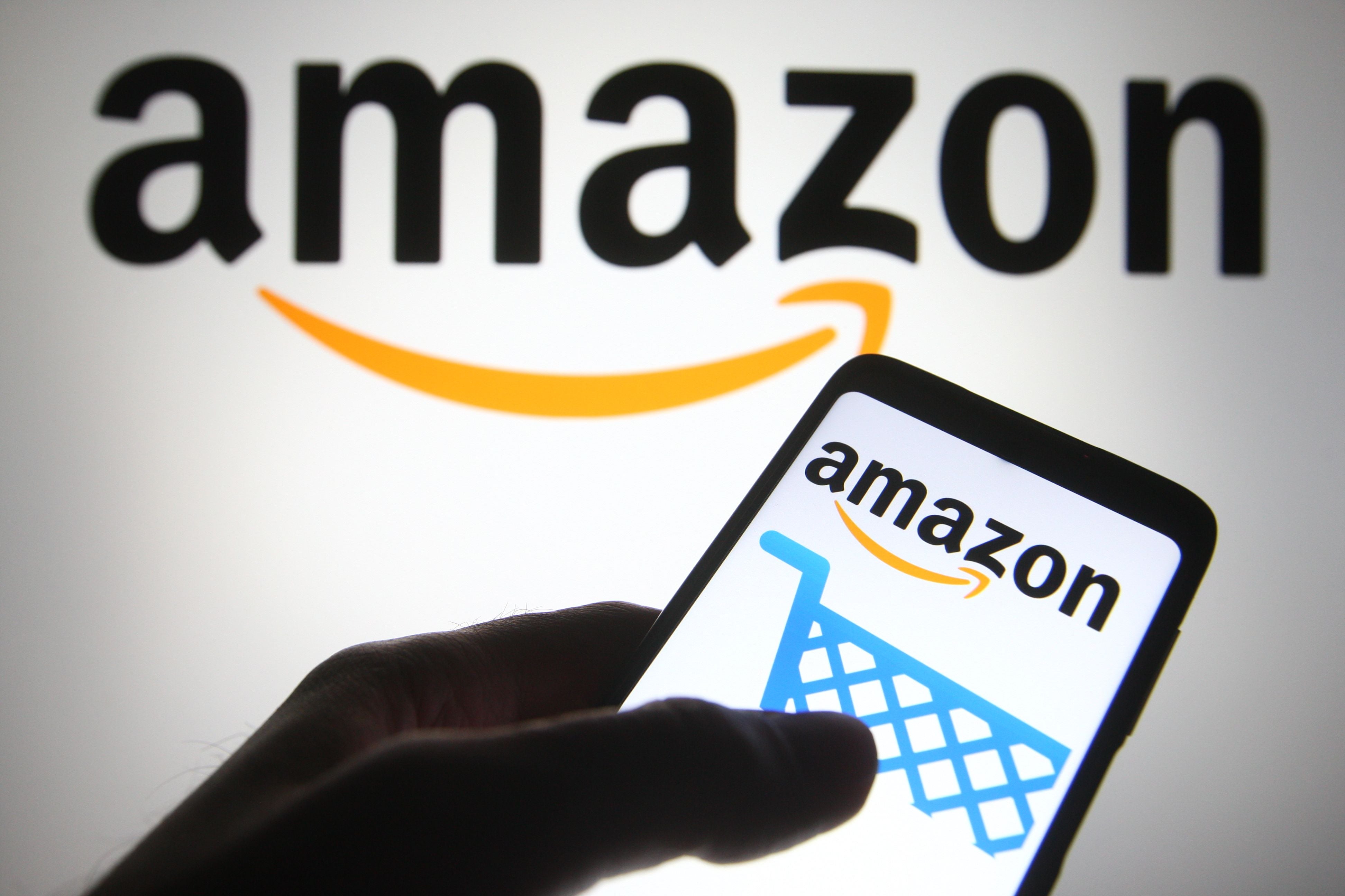 Amazon-Logo auf Smartphone und Bildschirm, Hand hält das Smartphone mit Einkaufswagen-Symbol.