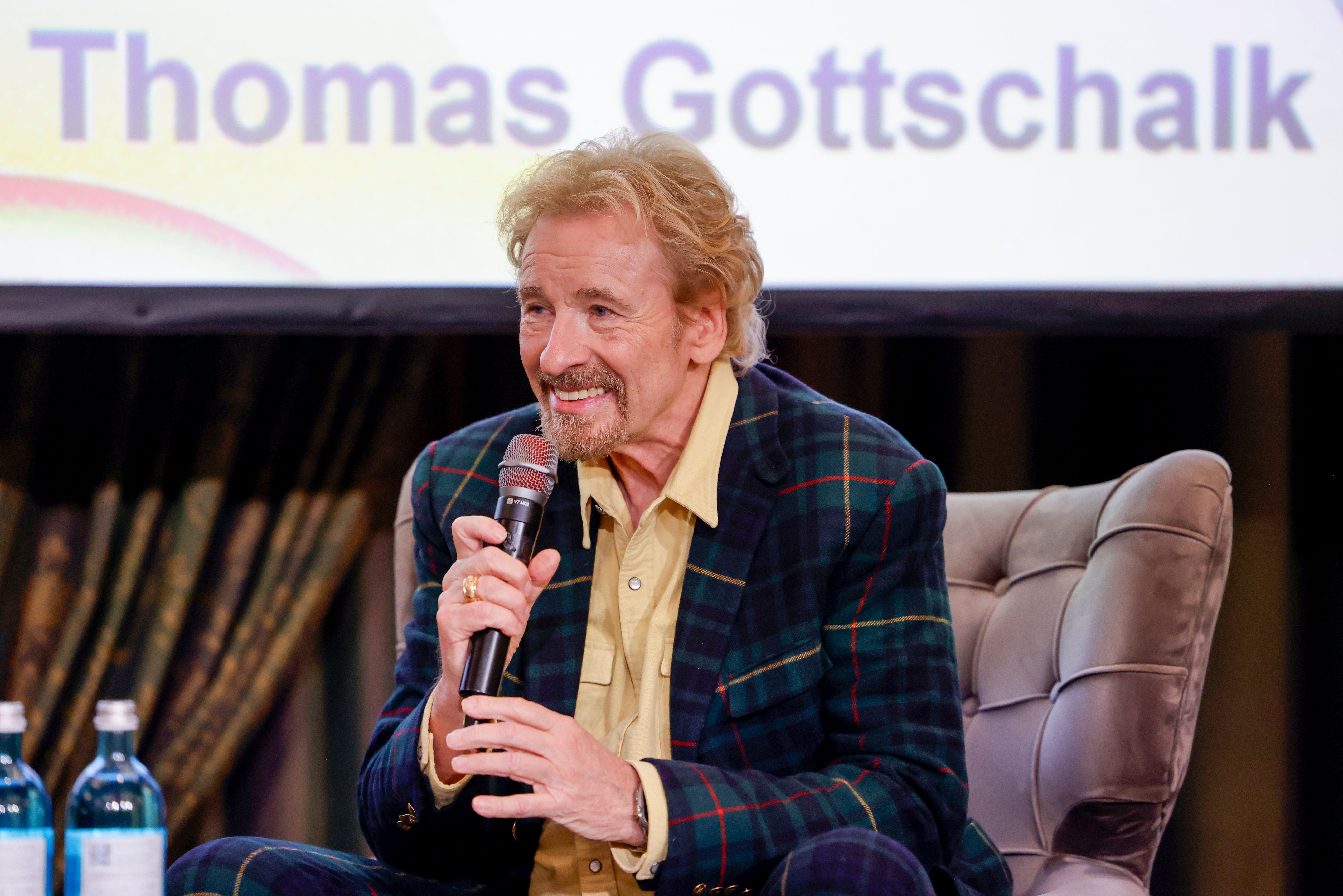 Ein Mann spricht mit Mikrofon auf einem Sessel, im Hintergrund steht "Thomas Gottschalk".