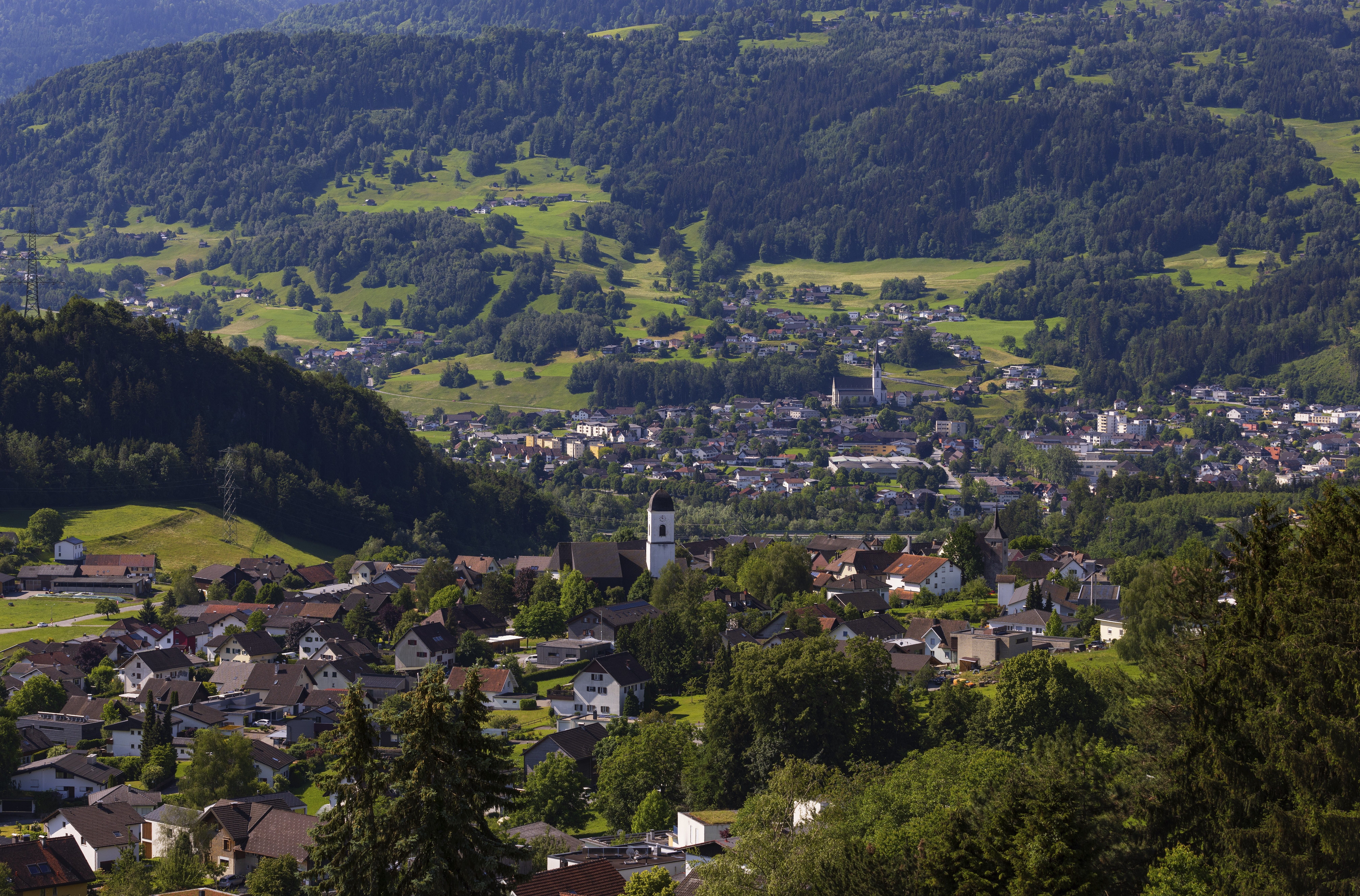 Blick auf ein Dorf mit Kirche, umgeben von grünen Hügeln und Wäldern in einer ländlichen Landschaft.