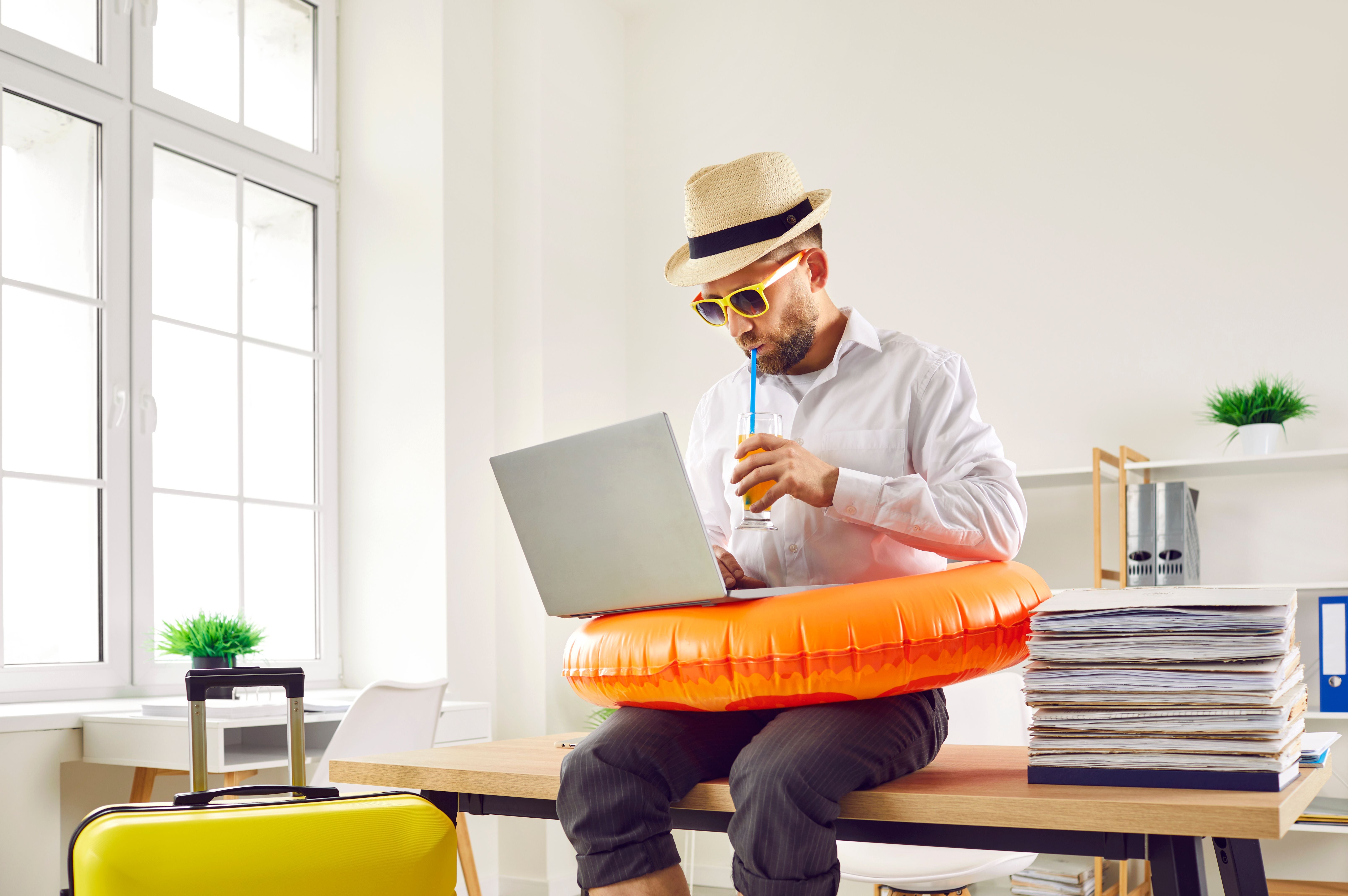 Mann im Büro trägt Sonnenhut und Schwimmring, arbeitet am Laptop und trinkt Orangensaft.
