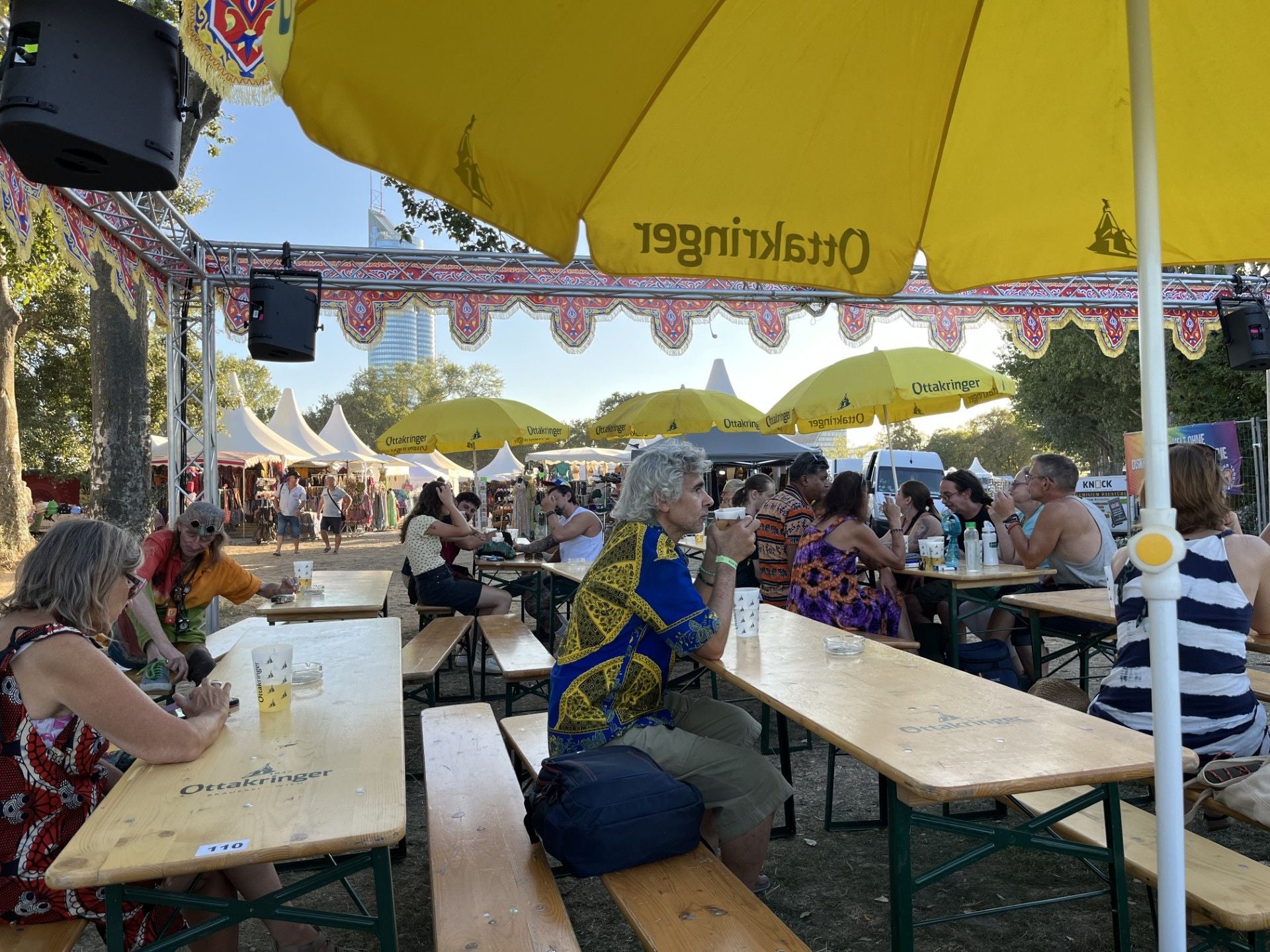 Menschen sitzen an Holzbänken unter gelben Sonnenschirmen bei Afrika Tage Festival im Freien.