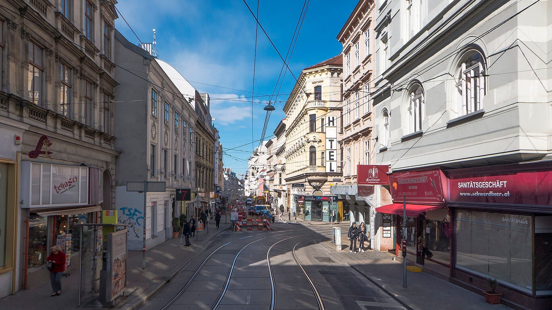 Straßenszene in der Neubaugasse mit Geschäften, Hotel und Straßenbahnschienen bei Tageslicht.