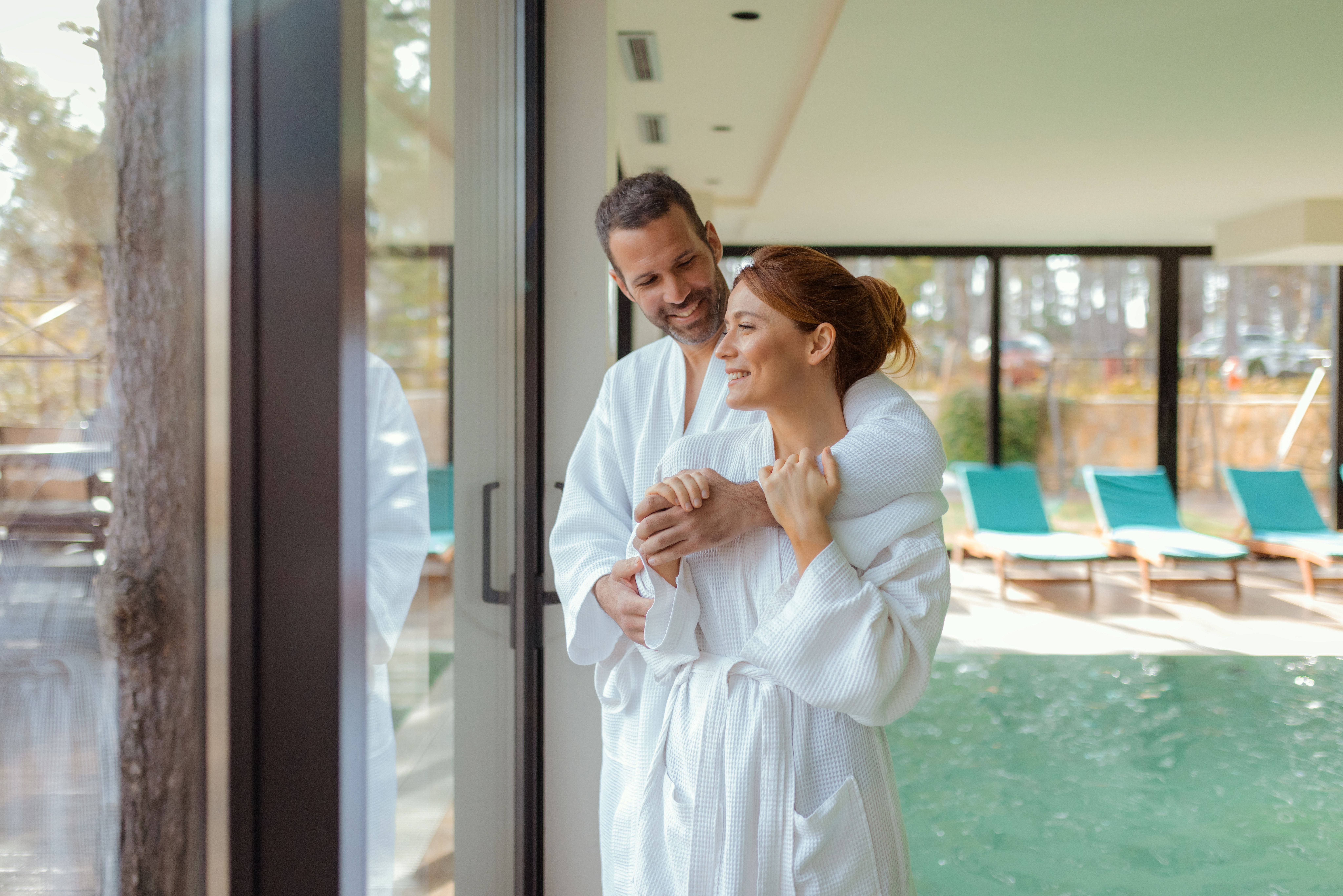 Ein Paar in Bademänteln umarmt sich lächelnd in einem modernen Spa am Fenster mit Pool und Naturblick.
