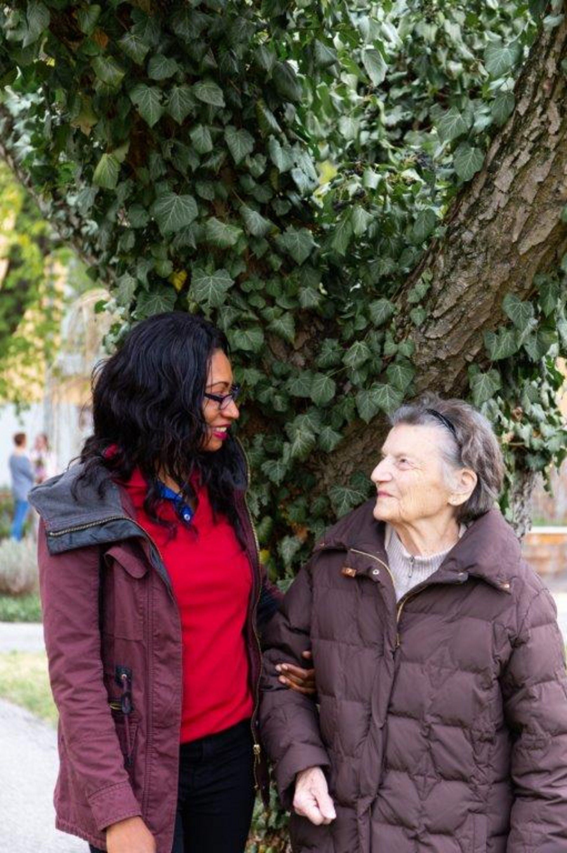 Junge Frau und ältere Dame stehen lächelnd nebeneinander vor einem Baum im Freien.