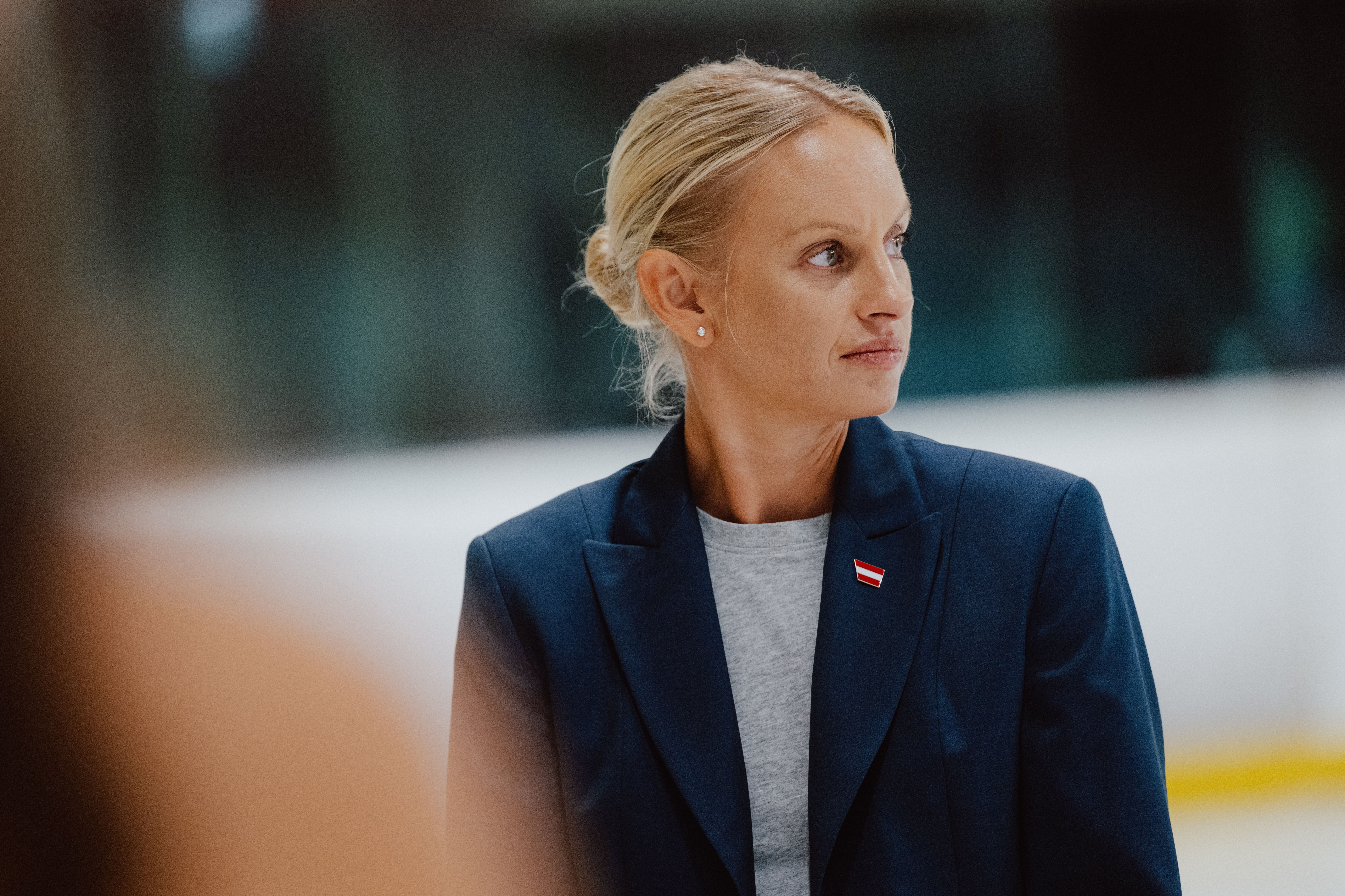 Frau im blauen Blazer mit Anstecker schaut zur Seite, unscharfer Hintergrund.