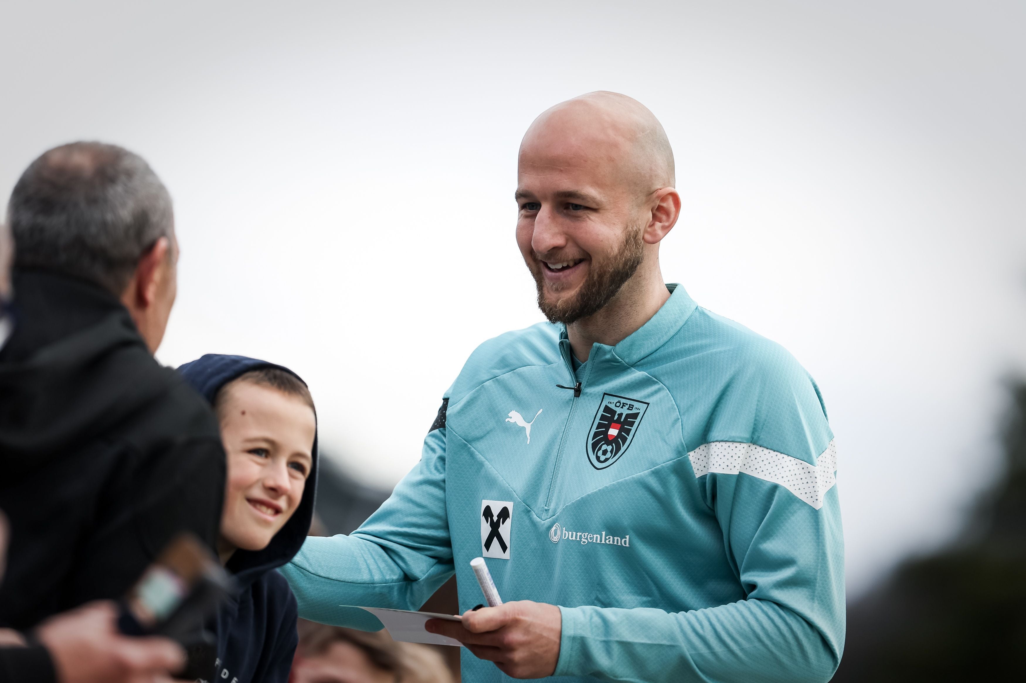Ein Fußballspieler im Trainingsanzug gibt Autogramme und lächelt mit Fans im Freien.