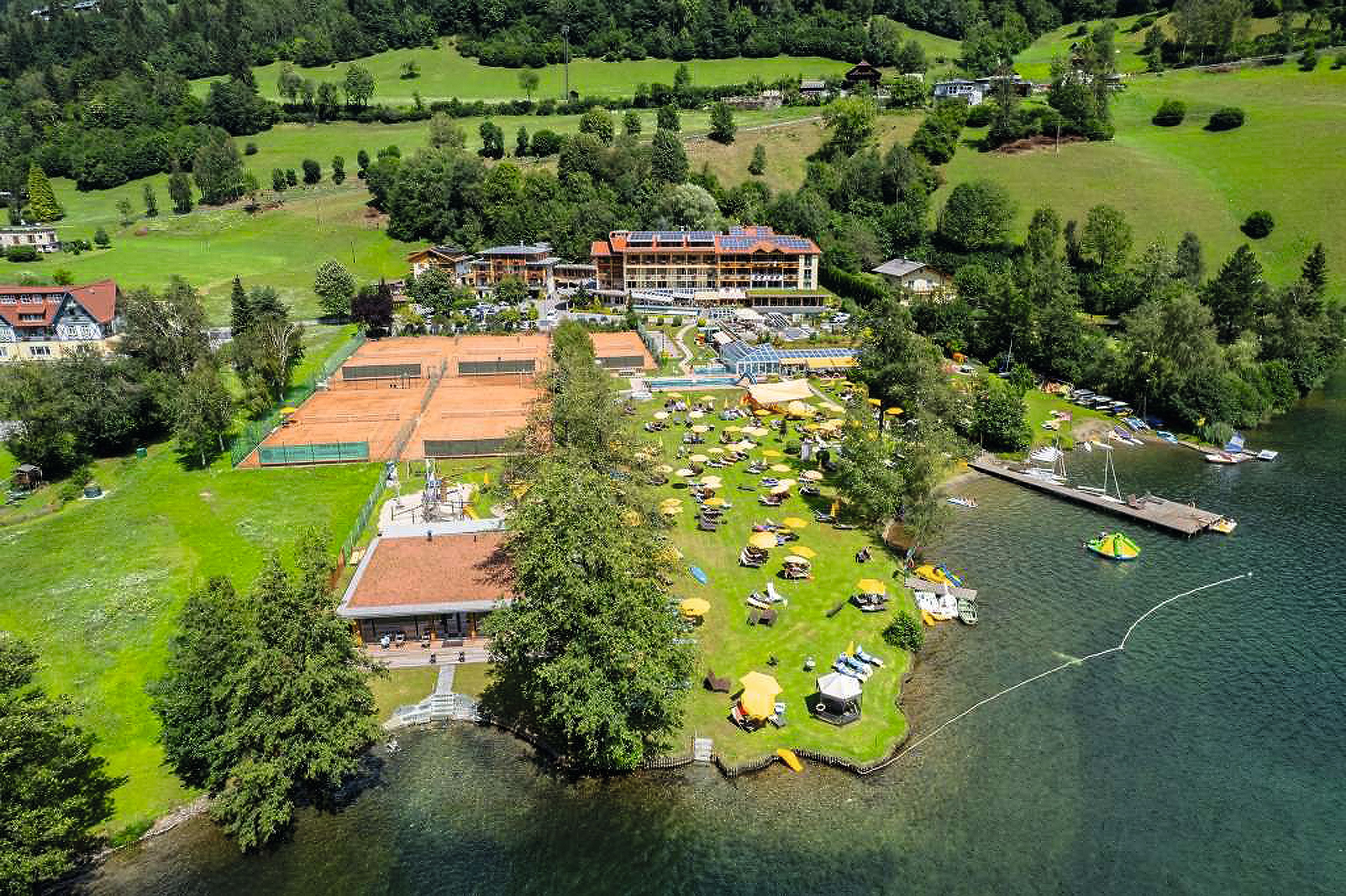 Luftaufnahme eines Hotels mit Tennisplätzen, Liegewiese und Steg an einem See in grüner Landschaft.