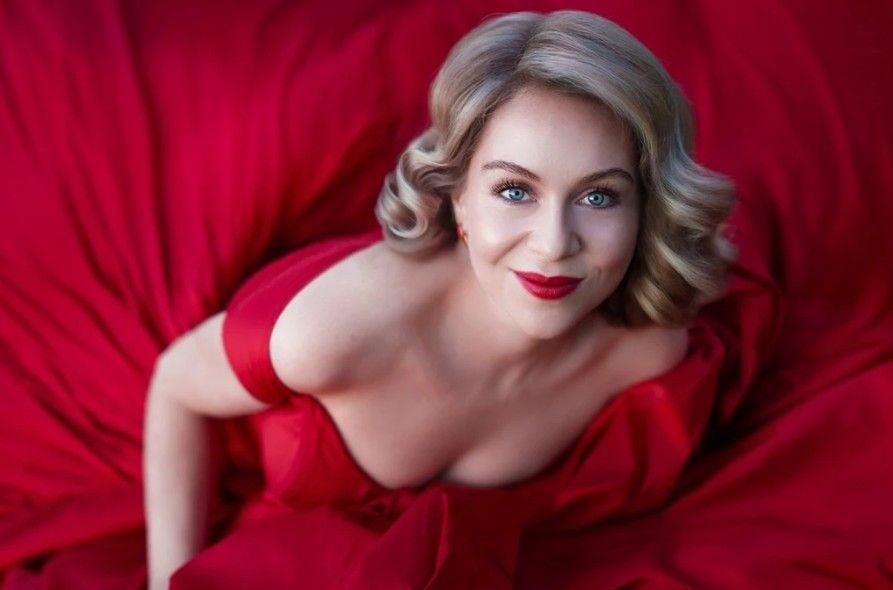 Frau mit blonden Locken in rotem Kleid lächelt in die Kamera, umgeben von rotem Stoff.