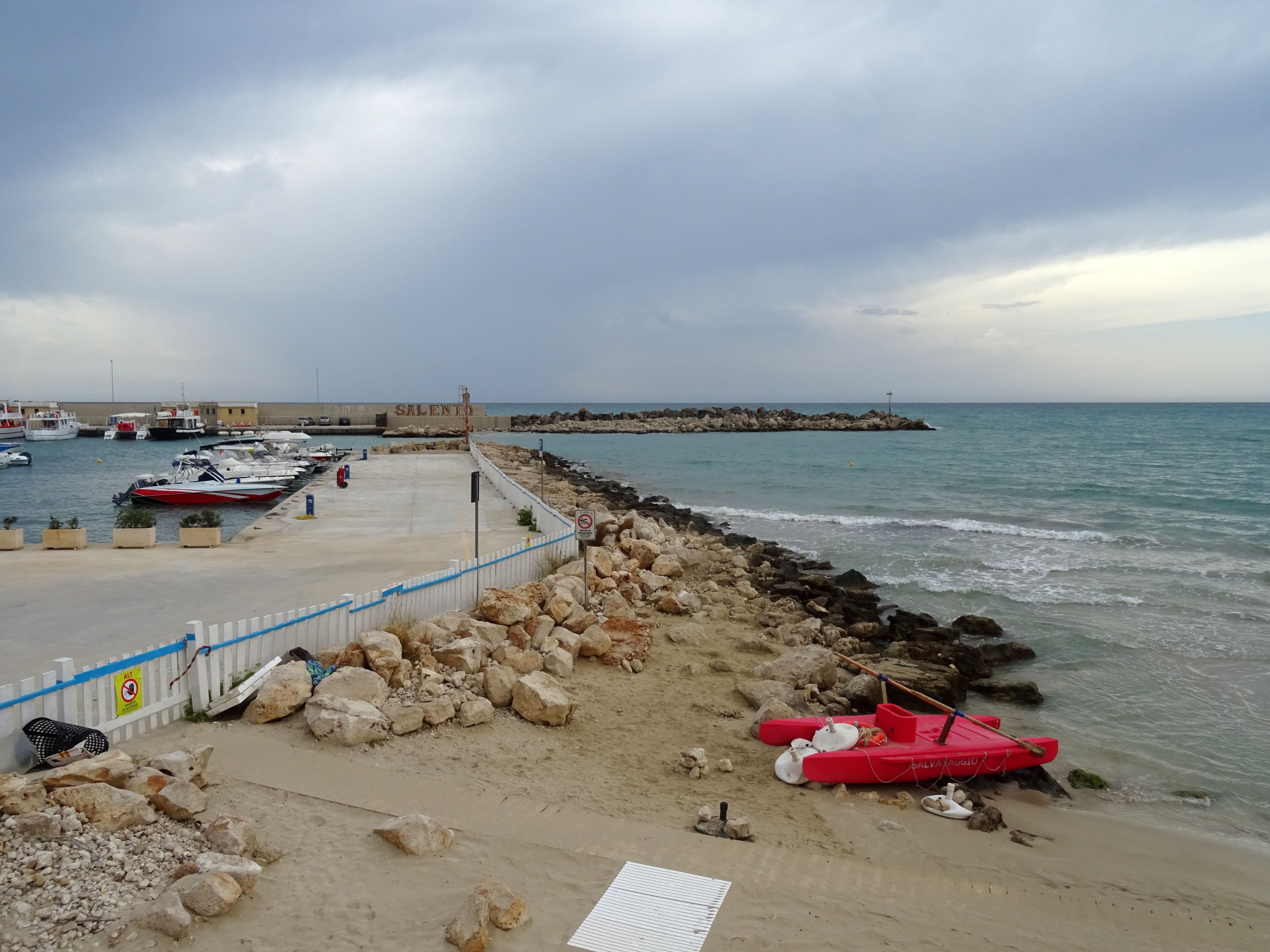 Blick auf einen kleinen Hafen mit Booten und einem roten Ruderboot am Sandstrand bei bewölktem Himmel.