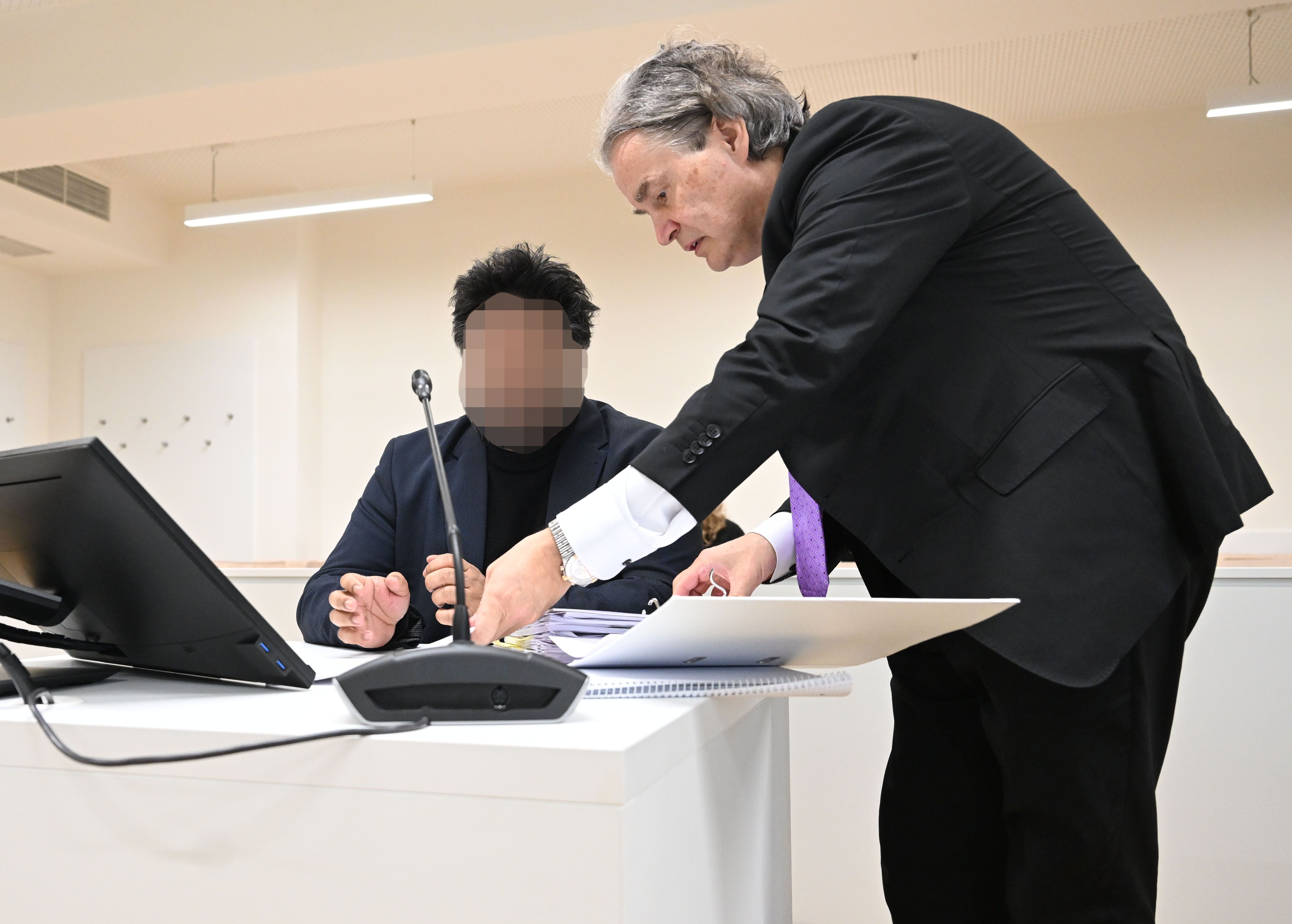 Ein Anwalt zeigt einem Mandanten in einem Gerichtssaal Unterlagen, beide sitzen an einem Tisch.