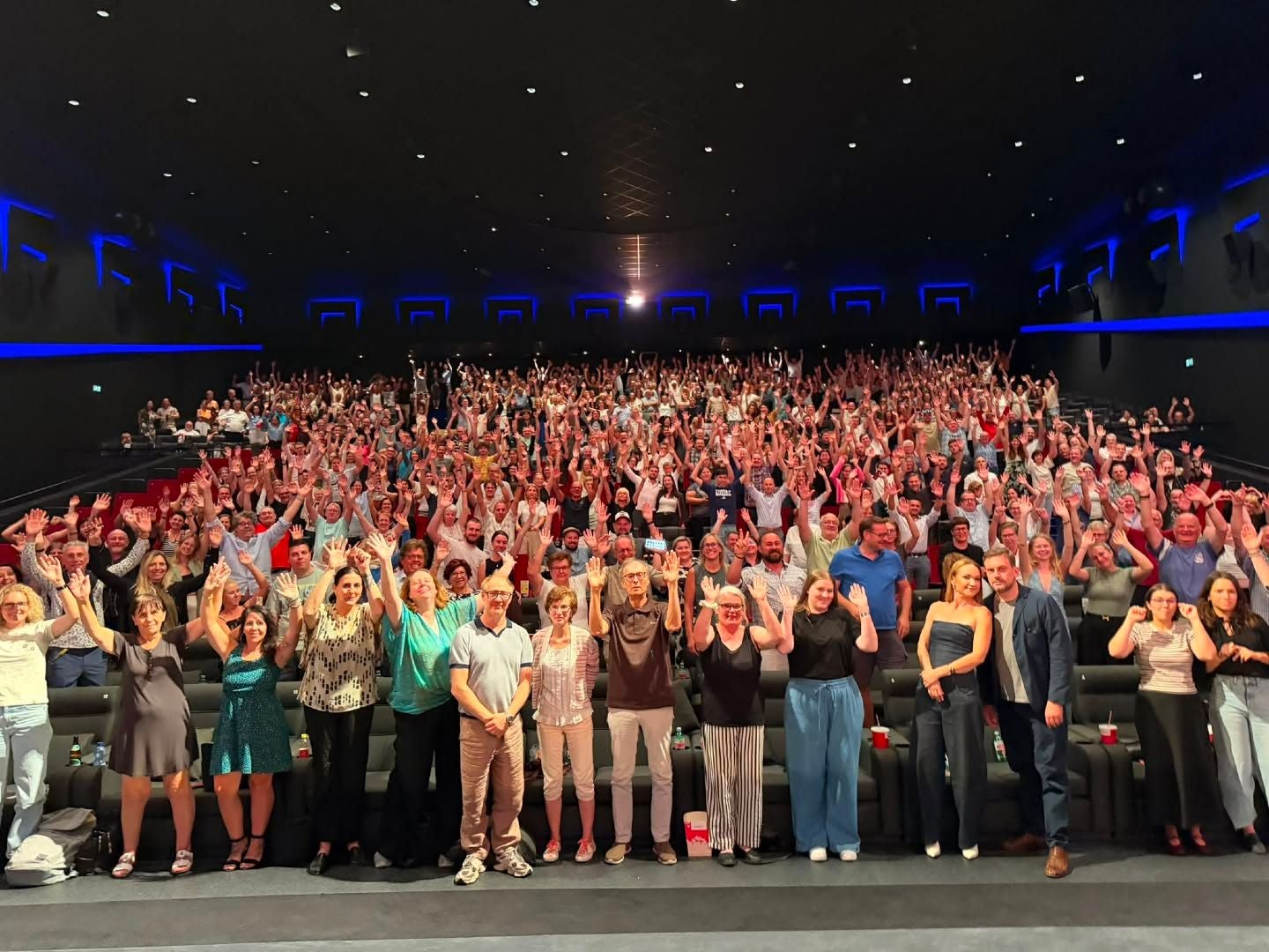 Große Menschenmenge in einem Kino, viele heben lächelnd die Arme, vorn eine Gruppe posiert.