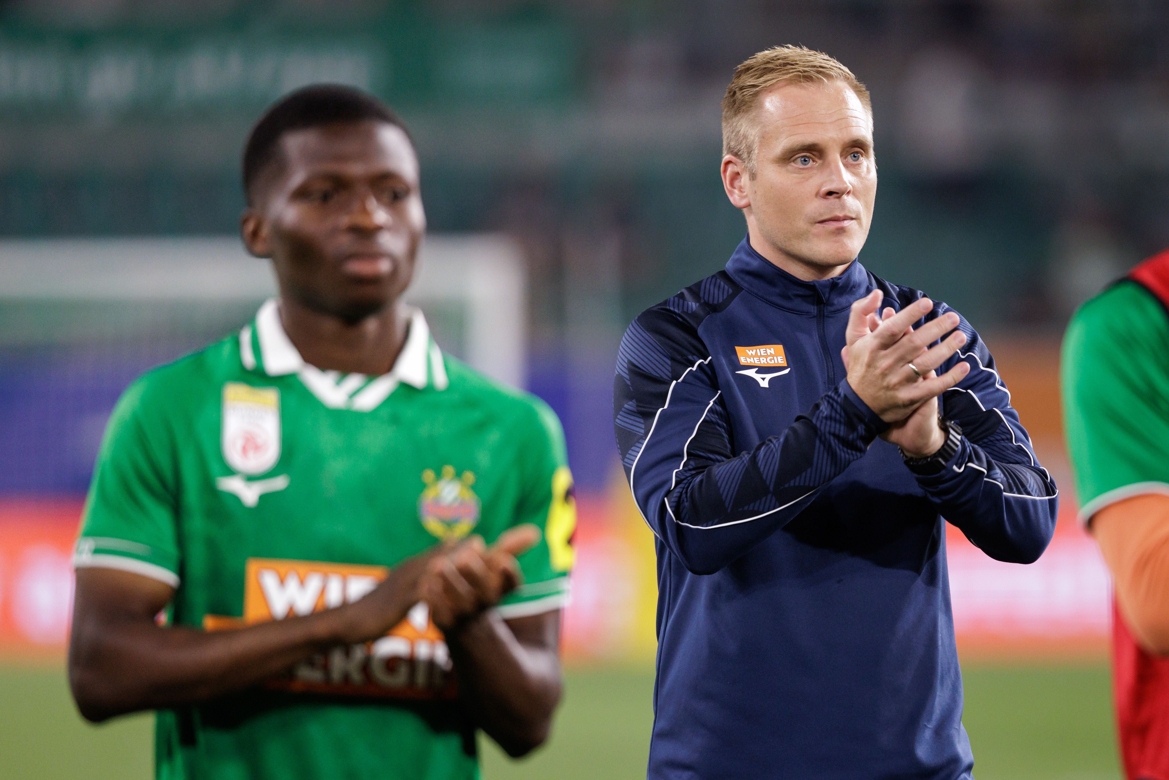 Zwei Männer im Rapid Wien Outfit klatschen nach einem Fußballspiel auf dem Spielfeld.