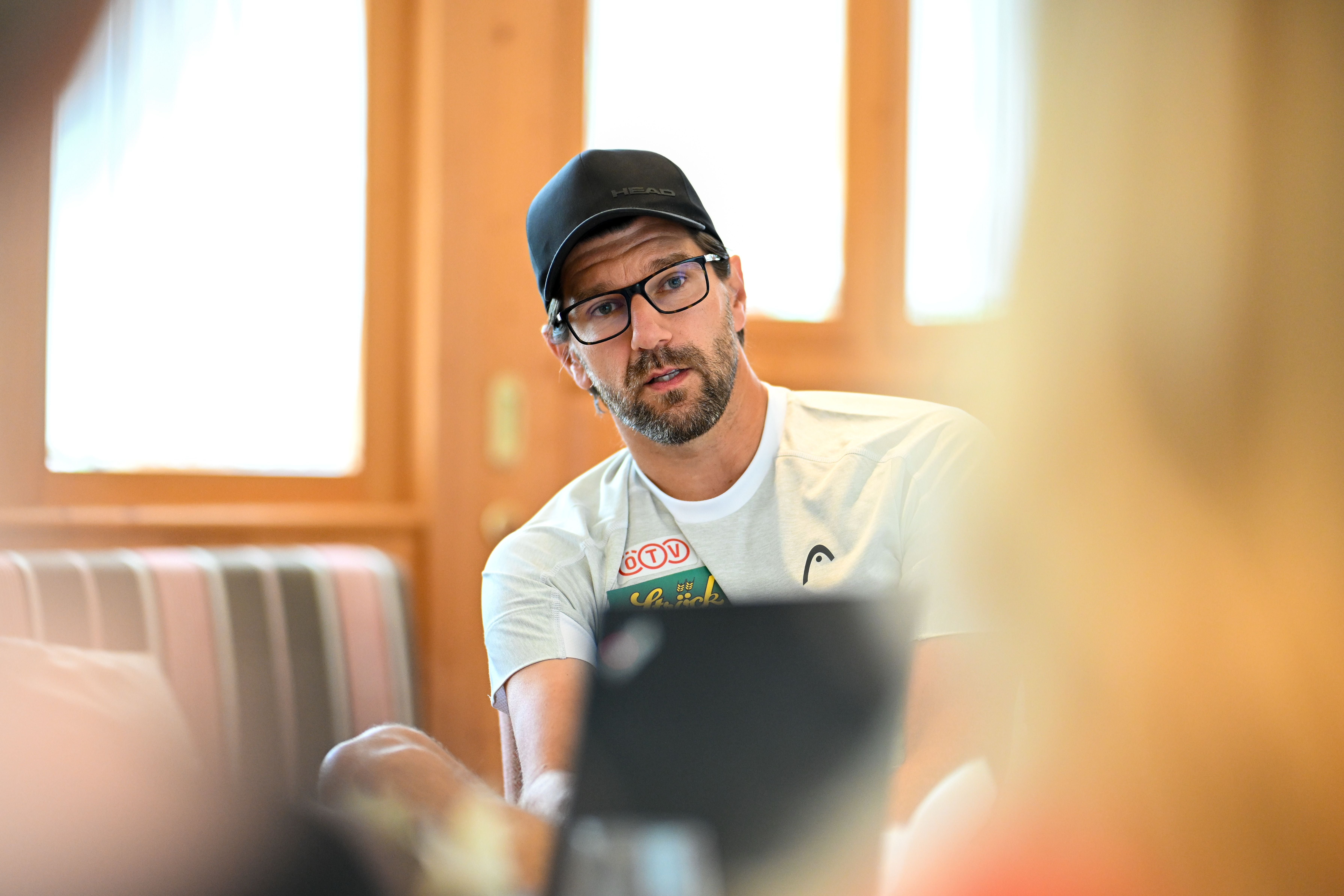 Ein Mann mit Brille und Kappe spricht bei einer Pressekonferenz in einem hellen Raum.