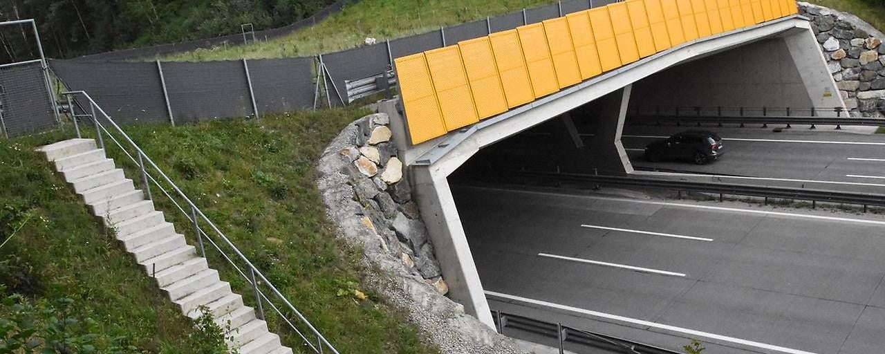 Straßenbrücke über Autobahn mit gelben Lärmschutzwänden, daneben eine Treppe am Hang.