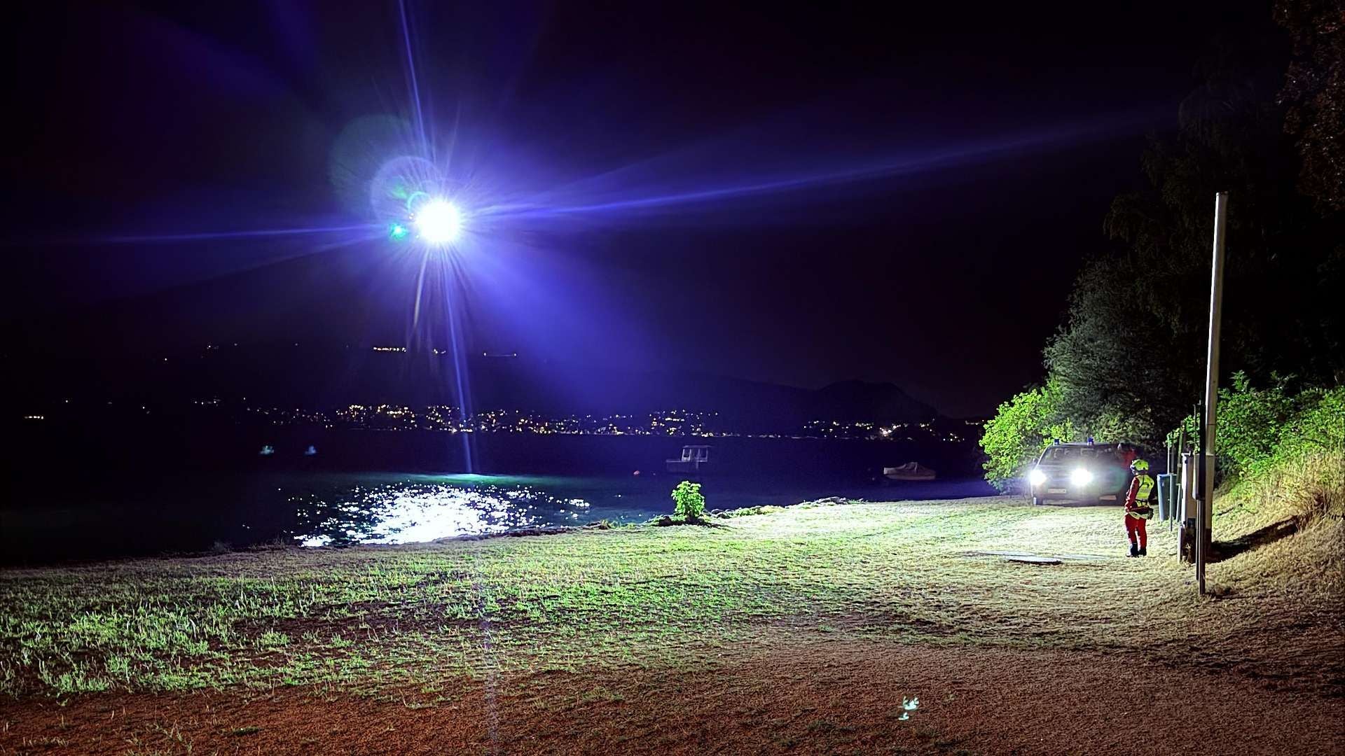 Ein Hubschrauber leuchtet nachts einen See aus, Rettungskraft steht am Ufer neben einem Auto.