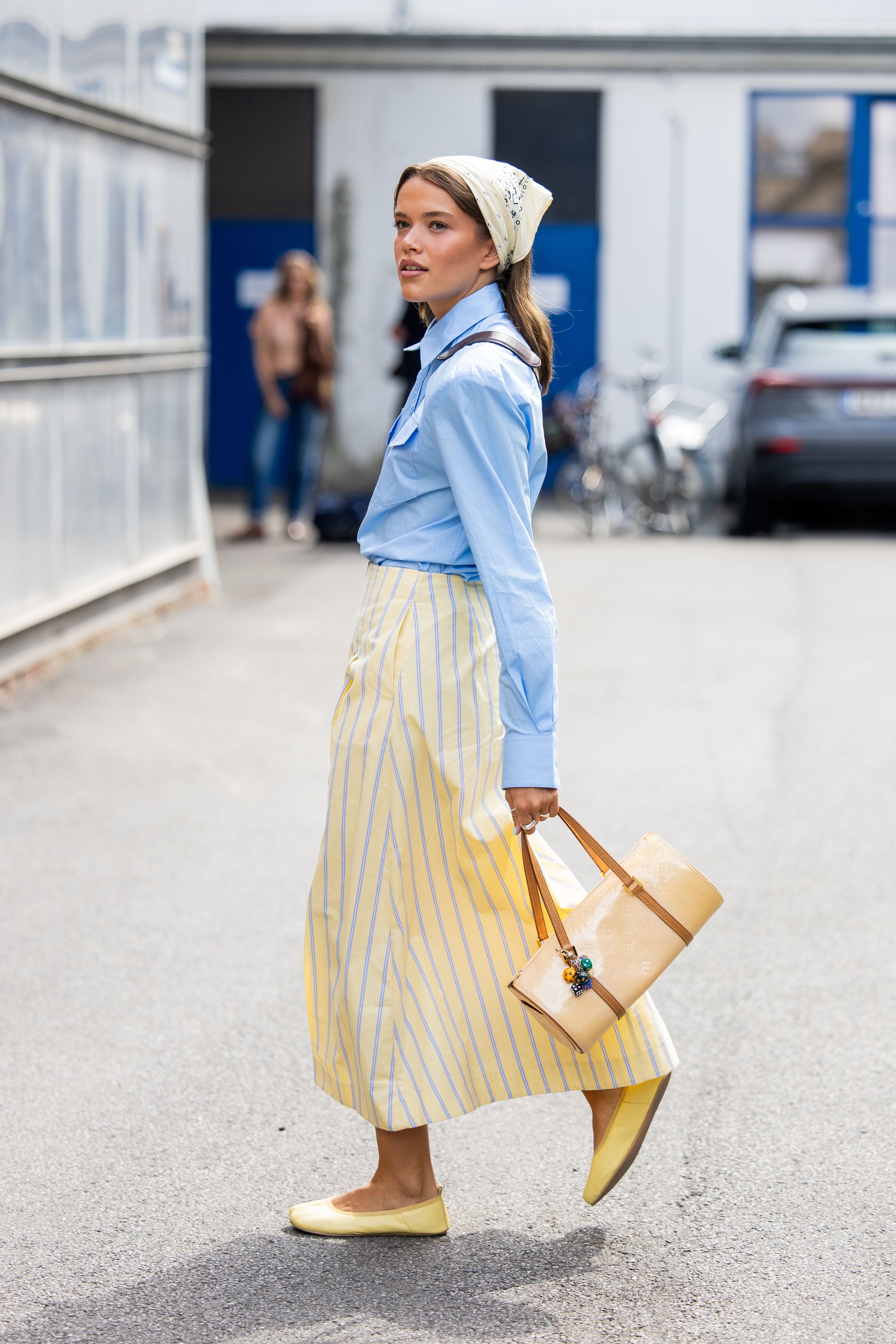 Frau mit hellblauer Bluse, gelbem Rock, Kopftuch und beiger Tasche auf der Straße.