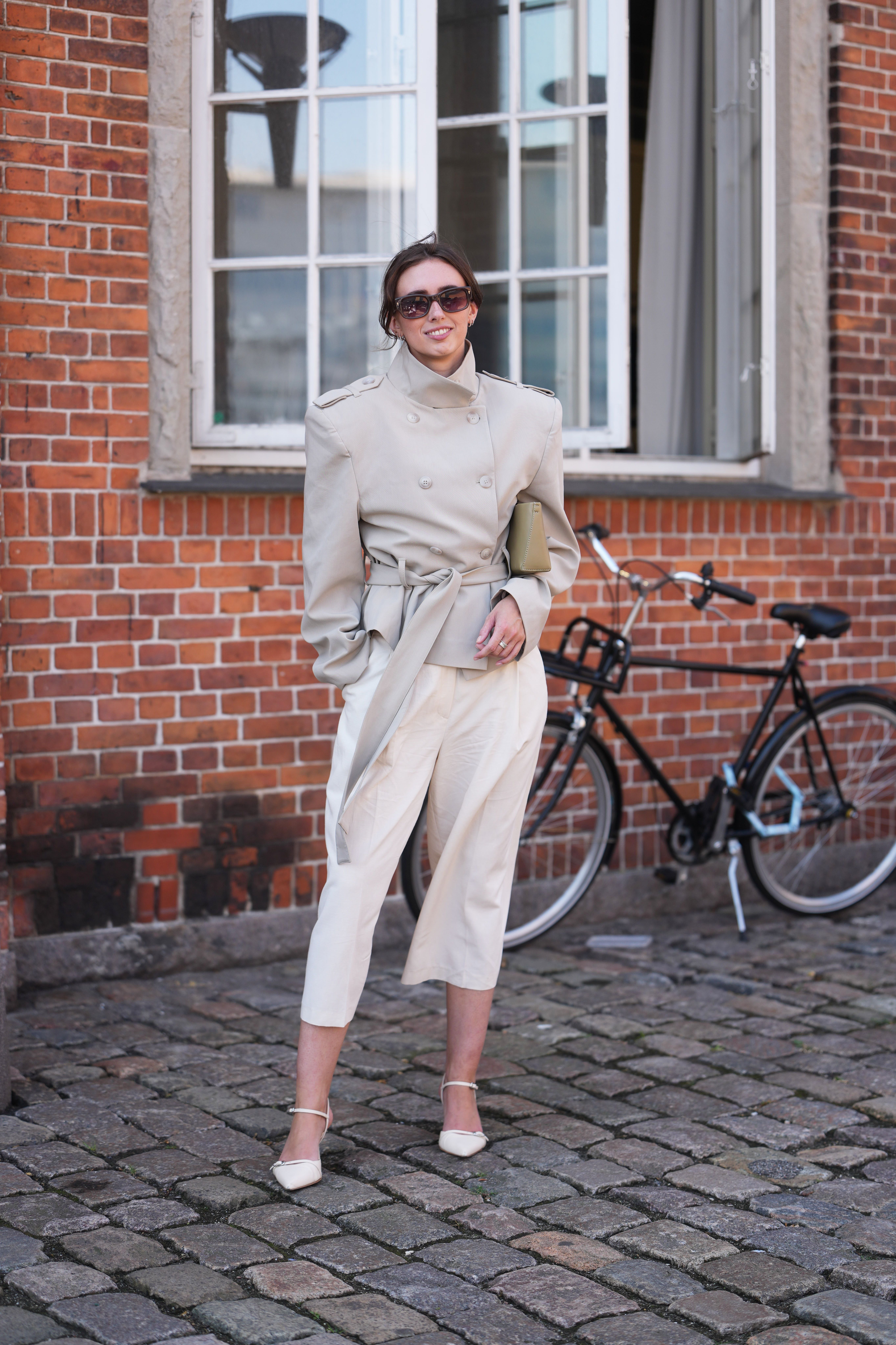 Frau mit beigem Trenchcoat, weißer Hose und Sonnenbrille steht vor einem Fahrrad und Backsteinwand.