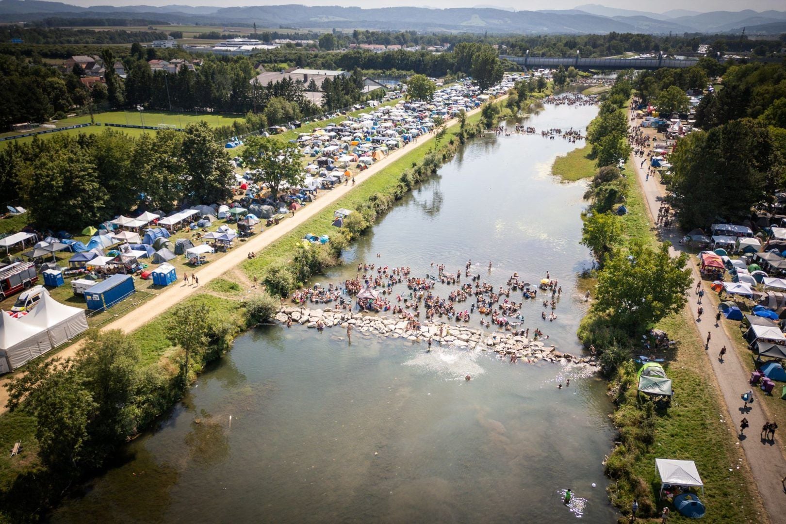 Luftaufnahme eines Flussufers mit vielen Zelten und Menschen, die im Wasser baden.